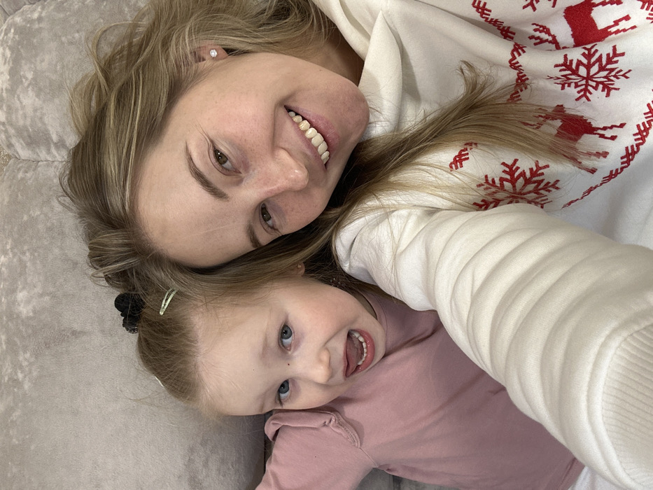 A woman and a young girl are lying on a couch. The woman has blonde hair, fair skin, and is wearing a white sweater with a red snowflake and reindeer pattern. She is smiling broadly, showing her teeth. The girl, with light blonde hair and blue eyes, also smiles. She wears a pink shirt and has a black hair clip in her hair. The camera is positioned overhead, capturing both faces in a close-up shot, emphasizing their joyful expressions.