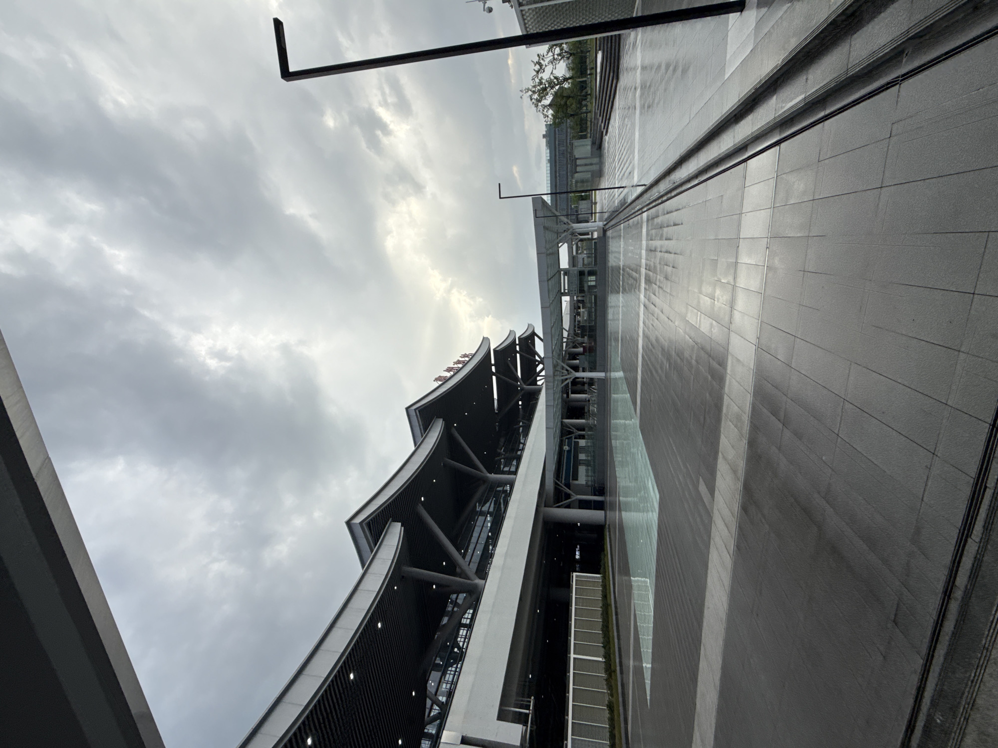 The image shows the exterior of a modern train station building, likely Huizhou North Railway Station, in Huizhou, China. The sky is overcast, with a mix of grey clouds and patches of brighter sky. The building features a distinctive curved roofline supported by large concrete pillars. The foreground is an expansive paved plaza, wet from rain, with reflective pools of water scattered across its surface. A dark, angular lamppost stands to the right of the frame. The overall scene is one of a modern transportation hub under a cloudy sky, with the wet pavement indicating recent rainfall. No people or specific actions are visible.