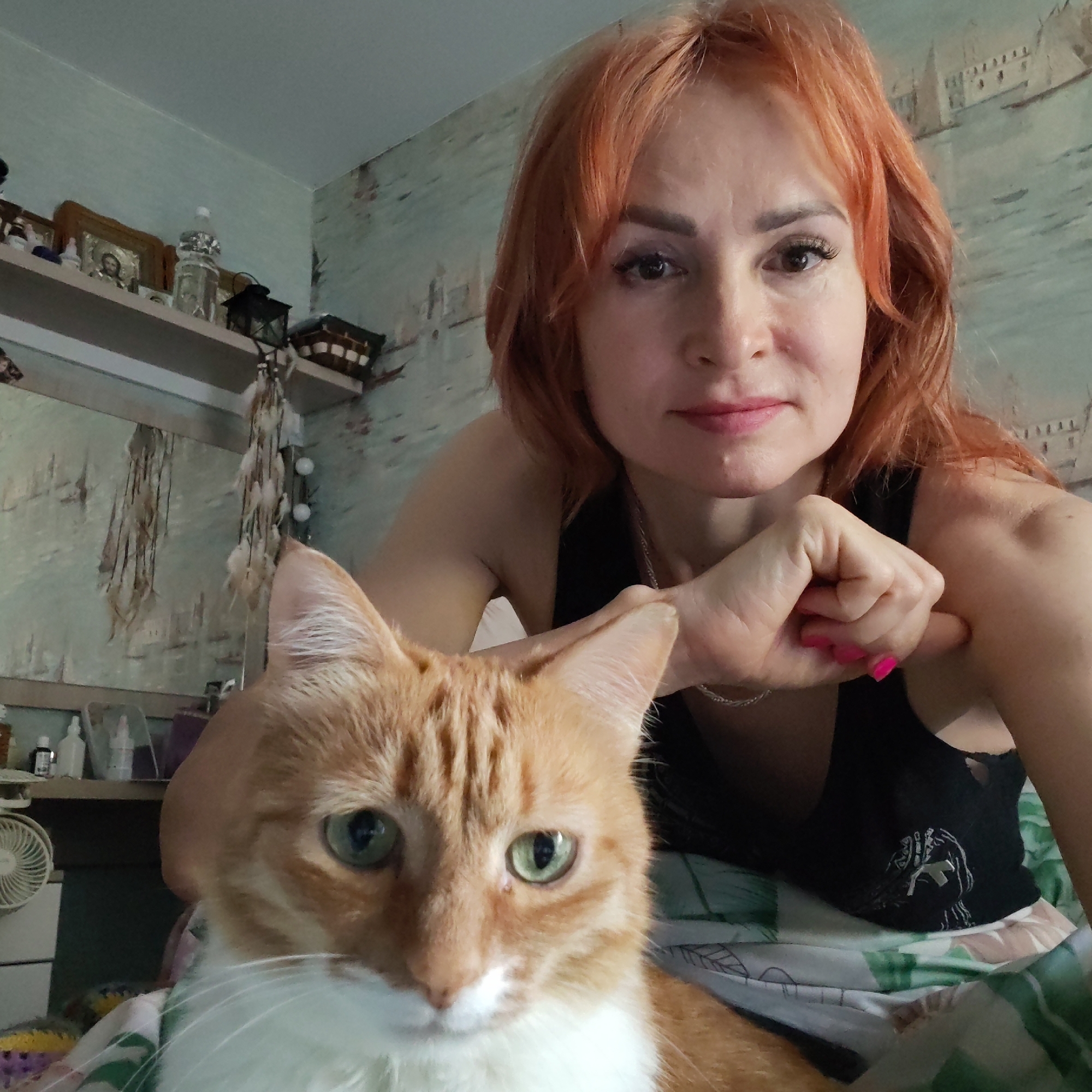 A woman with reddish-orange hair leans forward, looking into the camera with a slight smile. Her left hand is near her face, knuckles touching her neck, and her nails are painted pink. She's wearing a black tank top. An orange tabby cat with green eyes sits in the foreground, its face centered and looking forward. The background reveals a room with a shelf cluttered with objects. The wallpaper is a muted aqua color with a cityscape motif. To the left, a shelf holds small objects and decorations, and a white fan is visible. The location is likely Chaikovsky, Russia, based on the information provided.