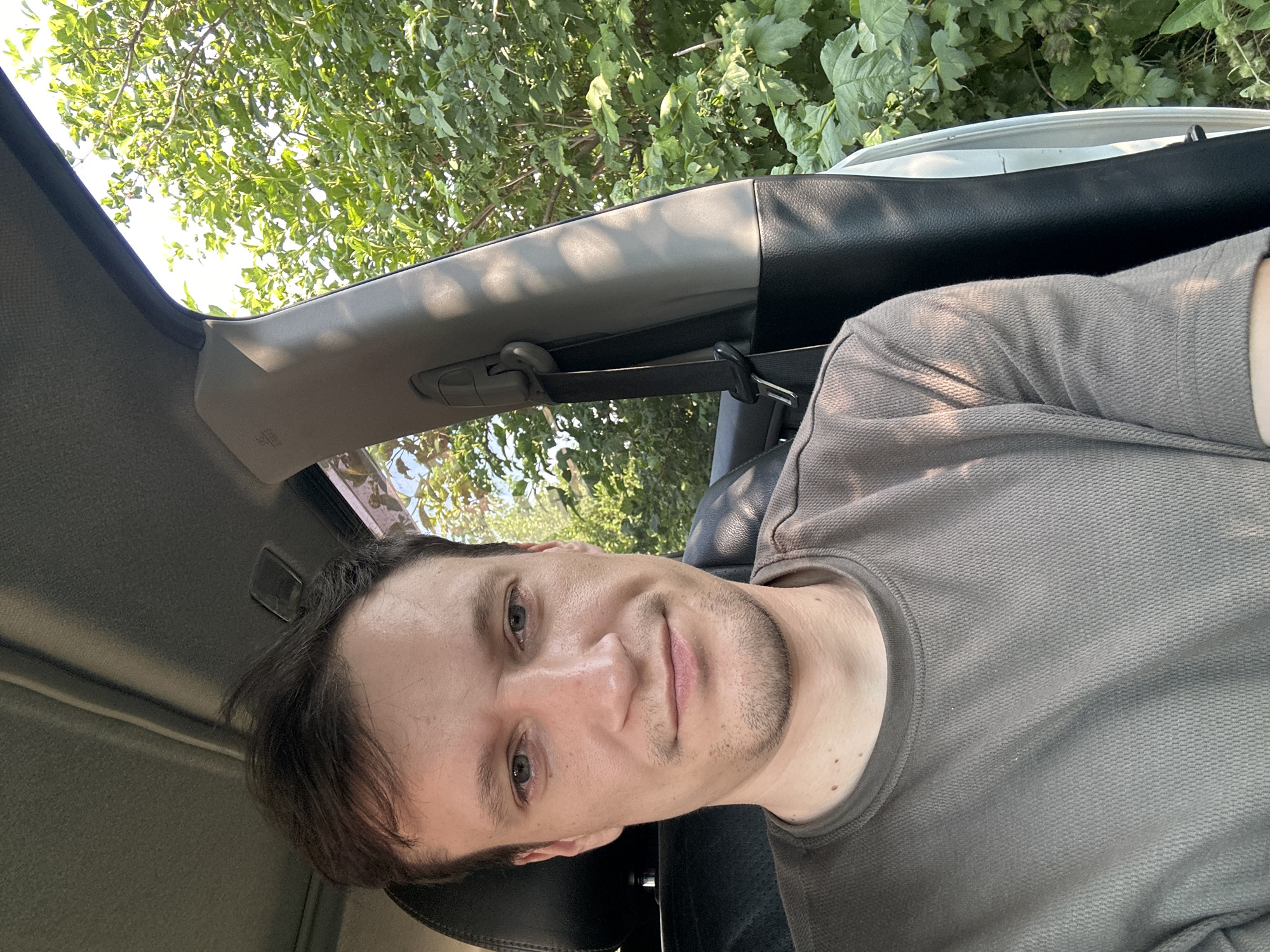 A man is taking a selfie inside a car. He is wearing a grey t-shirt and has short, dark hair. The car's interior is visible, including the seatbelt and a portion of the car door. Outside the car, lush green trees and foliage are visible, suggesting a natural or possibly rural setting. The image does not contain any information about the city of Shatava, Ukraine.