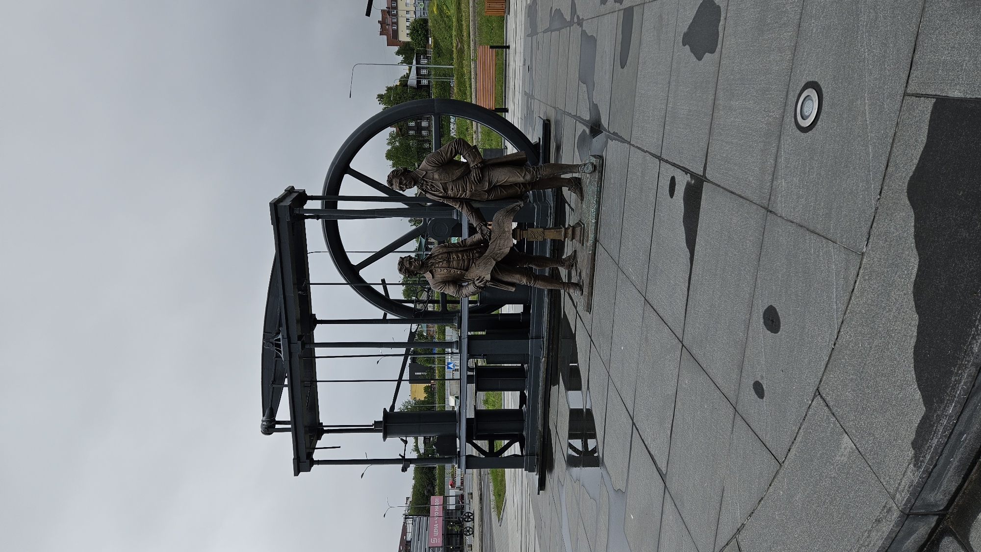The image captures a bronze sculpture group of three figures posed in front of a black mechanical structure with a large wheel. The figures are dressed in clothing reminiscent of the 18th century. The sculpture rests on a light gray tiled surface. The environment appears urban, as evidenced by distant buildings, streetlights, and greenery. The sky is overcast. The location is Unknown, Unknown.