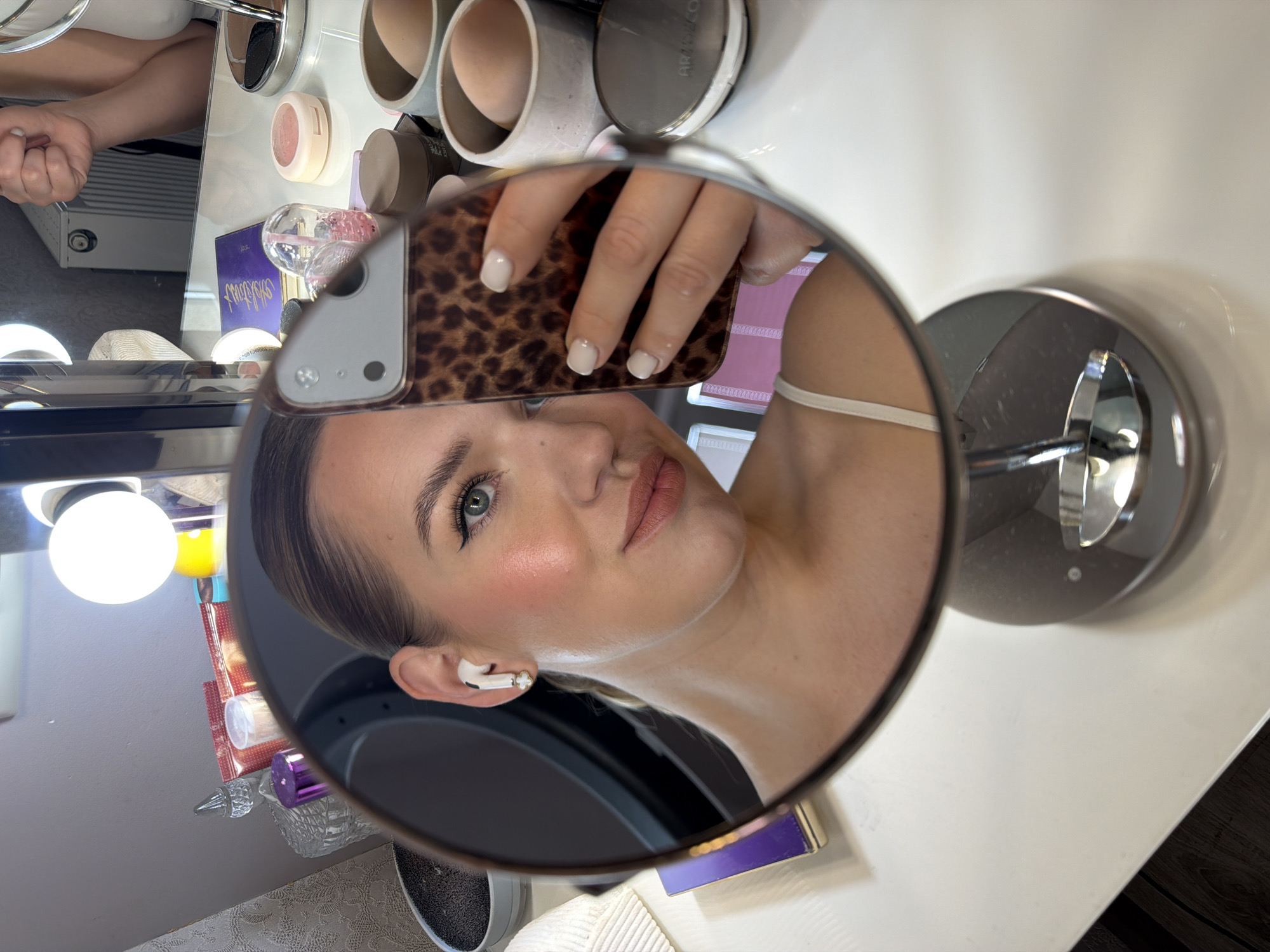 The image shows a woman taking a selfie in a vanity mirror. She is wearing a white top and has her hair pulled back. She is wearing white AirPods and has a makeup palette and blush on the table in front of her. The scene appears to be set up for applying makeup. The location is in Dreiliņi, Latvia.