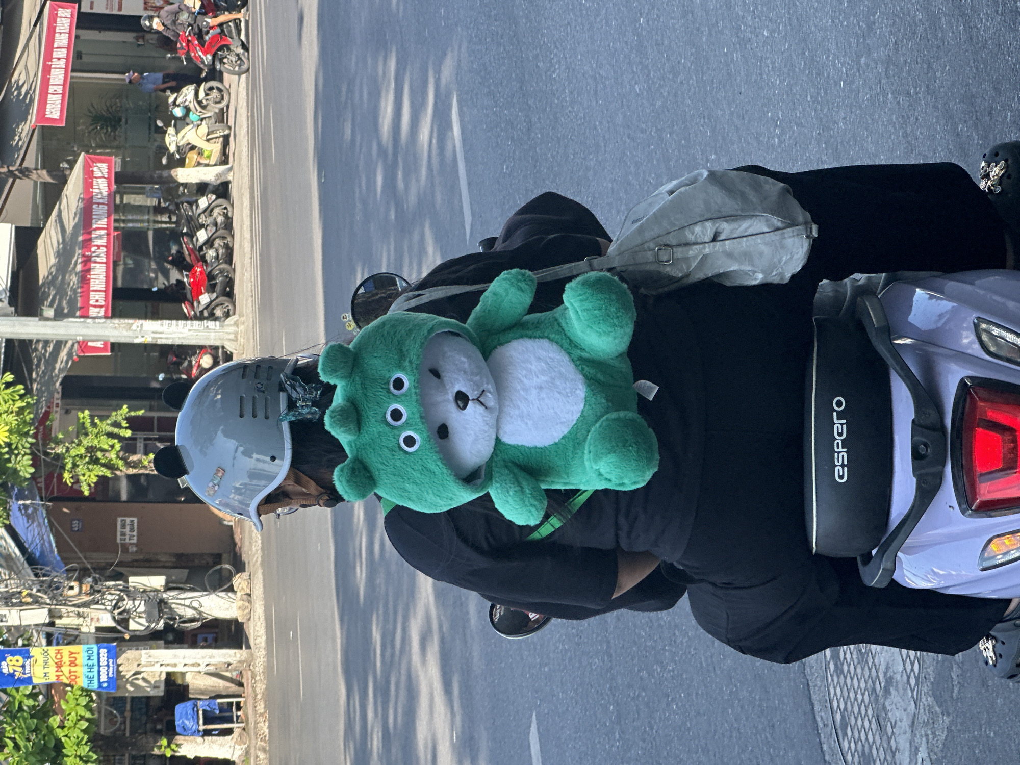 A person is riding a light purple scooter in Bac Nha Trang Ward, Vietnam. They are wearing a light grey helmet with black bear ears, a black t-shirt, and black pants. A green stuffed animal bear with three eyes is attached to the back of the helmet and backpack. A light grey messenger bag is slung over the person's shoulder. The scooter has the brand name "espero" on the seat. The background shows a street scene with buildings, trees, and other parked and moving motorcycles.