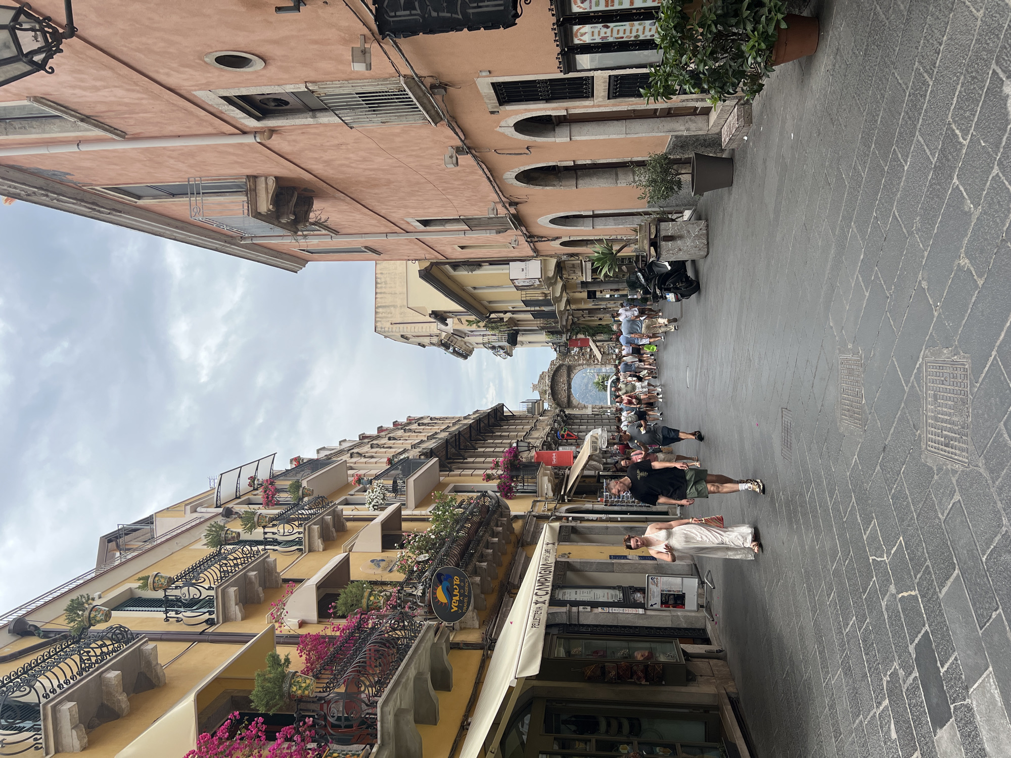 The image depicts a bustling street scene in Taormina, Italy, likely Corso Umberto. On the left, a yellow building with ornate balconies adorned with vibrant pink flowers and potted plants lines the street. Shops with awnings are visible on the ground floor. Several people are walking along the cobblestone street. A couple in the foreground, a woman in a white dress and a man in a black t-shirt and shorts, are walking towards the viewer. Further down the street, a crowd of people are visible, moving towards an arched gateway in the distance. The building on the right is painted a peachy-pink color and also features balconies. A black scooter is parked on the right side of the street. The sky is overcast.