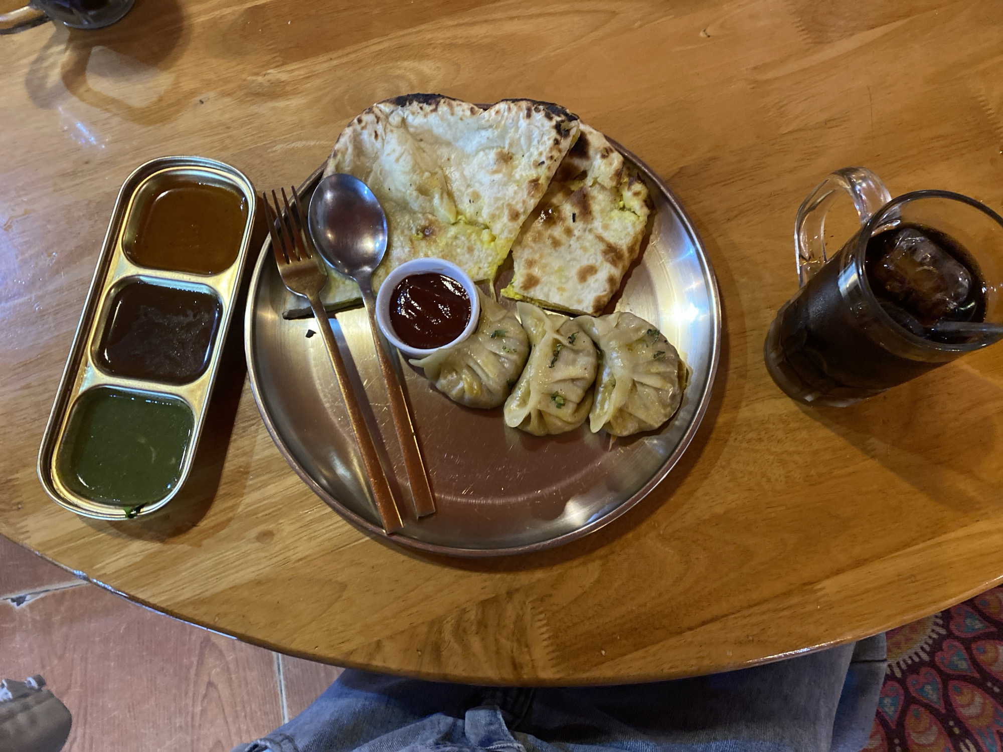 An overhead shot captures a meal on a wooden table. A stainless steel plate holds three momos, a small bowl of red sauce, and two pieces of naan bread, one folded over the other. To the right of the plate, a glass mug filled with a dark beverage and ice rests on its side. Above the plate, a rectangular metal tray contains three small compartments, each filled with a different dipping sauce: green, dark brown, and reddish-brown. A fork and spoon are laid across the plate. Part of a person wearing denim jeans is visible in the lower left corner. The background shows a portion of the wooden table and a glimpse of patterned fabric on the floor.