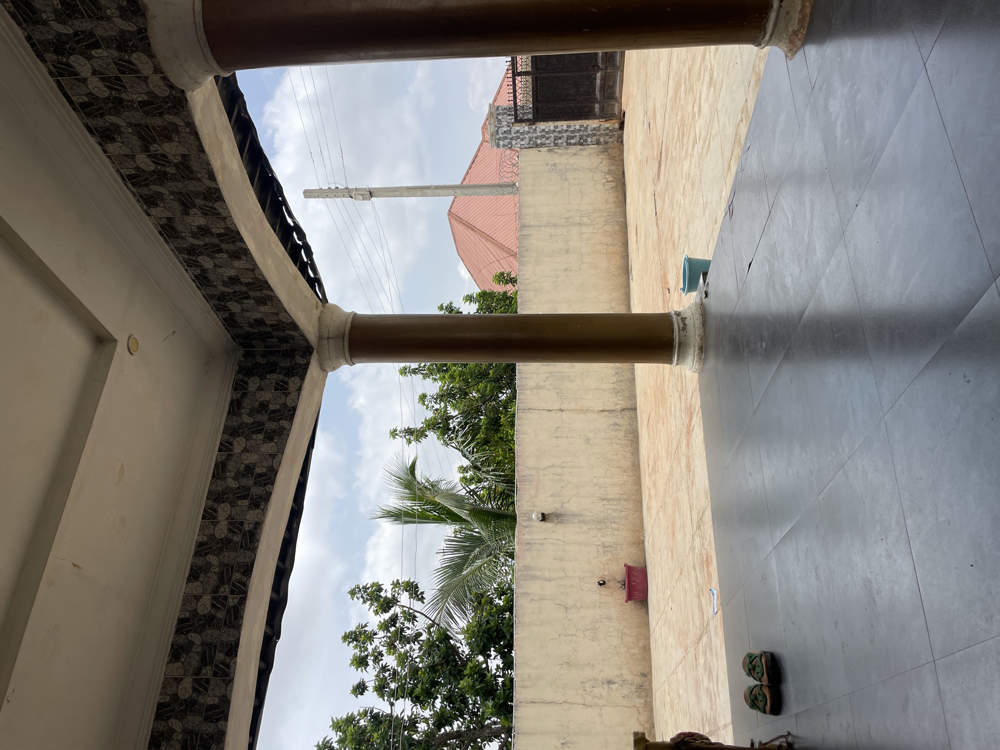 The image depicts a view from under a porch in Rafin Bauna, Nigeria. The porch is supported by two brown columns with decorative tops and bases. The porch ceiling is white with a decorative tiled border. Outside the porch, there is a tan-colored concrete wall with a small red flower pot attached. Beyond the wall, there are trees, a house with a red roof, and a black metal gate. The ground under the porch is covered with dark gray tiles, while the ground outside the porch is light tan. A pair of green sandals is on the ground under the porch. Power lines run across the sky.