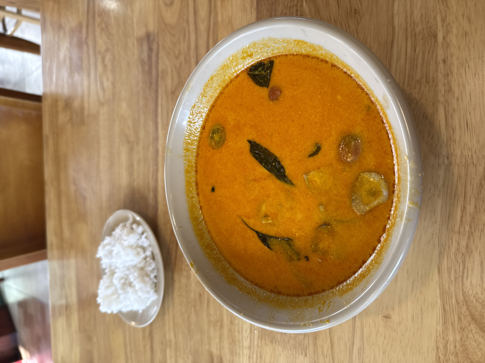 A bowl of orange curry sits on a wooden table next to a small dish of white rice. The curry appears to contain herbs, cherry tomatoes, and possibly tofu. The scene is likely in a restaurant within Nha Trang Ward, Vietnam, implied by the image context. No people are visible, and the focus is solely on the food arrangement.