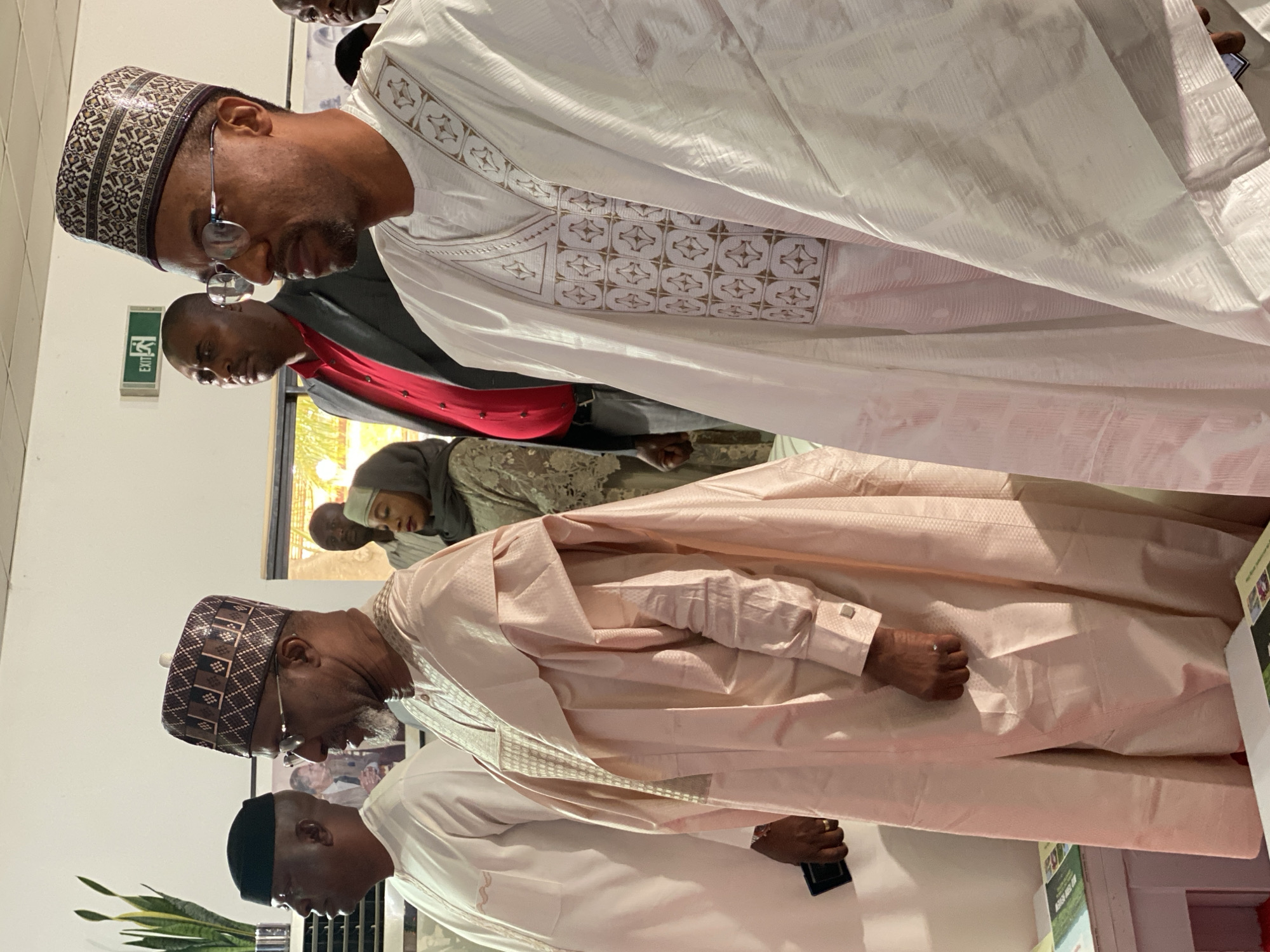 The image depicts a group of people indoors, likely in Kwarau, Nigeria. Several men are prominently featured, dressed in traditional Northern Nigerian attire, including elaborately embroidered gowns and caps. One man wears a white gown with detailed geometric patterns, while another sports a light pink gown. Some of the men wear glasses and are looking down. A woman in a head covering is partially visible in the background. An emergency exit sign is visible on the wall. The environment appears to be well-lit, possibly a meeting or gathering space. A man is seen holding what appears to be a cellphone. A stack of papers or leaflets are visible.