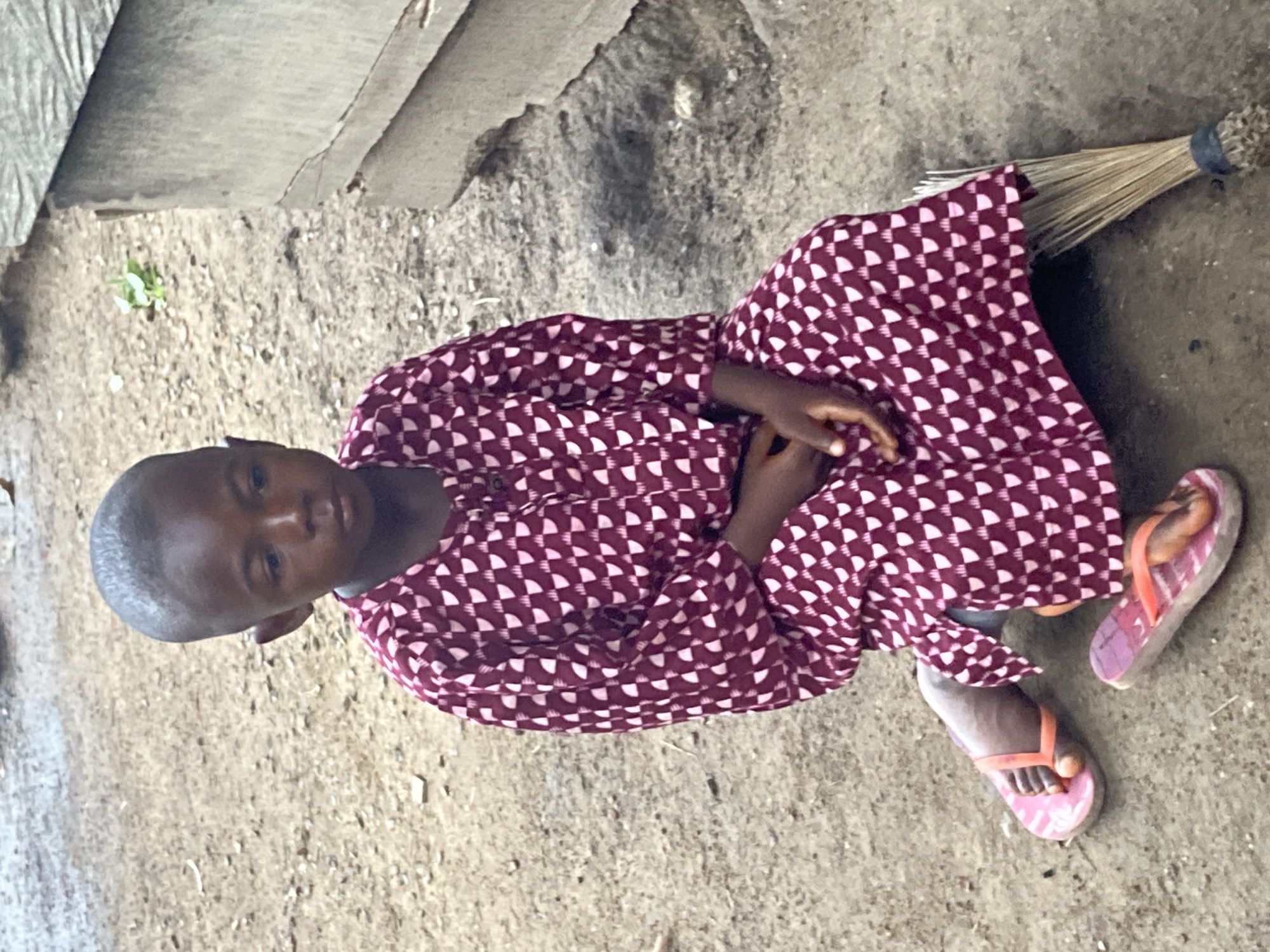 A young child is seated outdoors in Mararaba, Nigeria. The child, wearing a patterned maroon and white long-sleeved garment and pink flip-flops, is sitting on a dirt or concrete surface. A broom lies near the child. The background includes a concrete or stone structure and the ground.