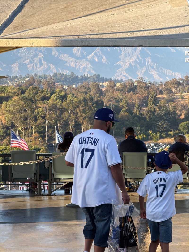 The image shows a father and son wearing "Ohtani 17" jerseys at Dodger Stadium in Los Angeles, United States. The view from the stadium includes trees and mountains in the background. The American flag is visible to the left.