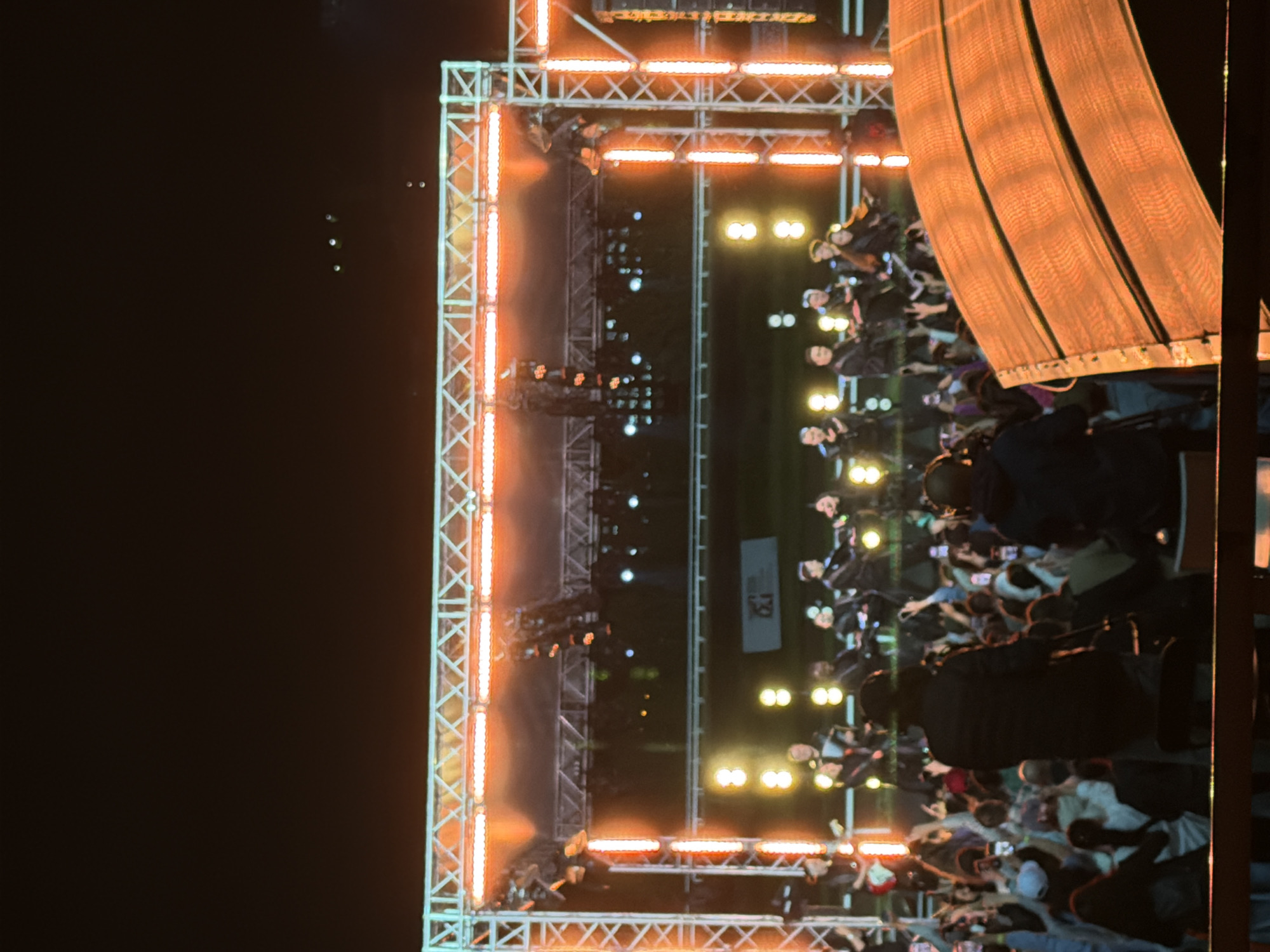An outdoor concert is in progress at night, with a stage set up and illuminated by bright orange lights. A group of performers, likely musicians, are on stage playing instruments. In front of the stage, a large crowd of people is gathered, many of whom are holding up their phones to capture the event. The lighting creates a vibrant and energetic atmosphere, with the orange glow reflecting off the stage equipment and the faces of the audience. The background is dark, suggesting it is nighttime, and some distant lights are visible, hinting at the urban environment. The scene depicts a lively concert experience with performers engaging their audience.