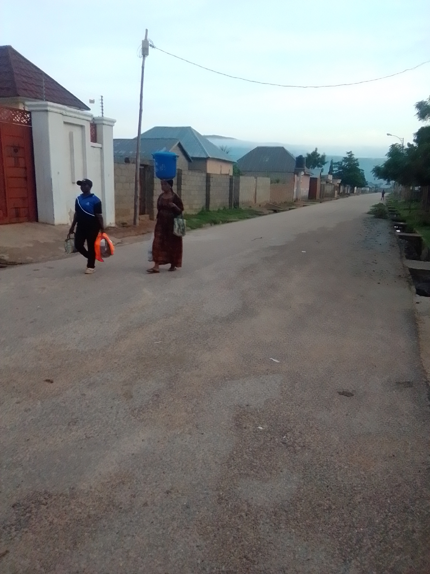 In Liji Hill, Nigeria, at dusk, two individuals walk along a paved road lined with single-story buildings and some vegetation. On the left, a person wearing a dark baseball cap, a blue and black shirt, and dark pants walks away from the camera, carrying a white bag in their left hand and an orange object in their right. To their right, a woman with dark hair wears a long, patterned brown dress and a blue bucket on her head. She carries a lighter-colored bag over her left shoulder. The road is empty except for these two individuals and a distant figure on the right side of the road. The sky is overcast, and the light suggests the end of the day.