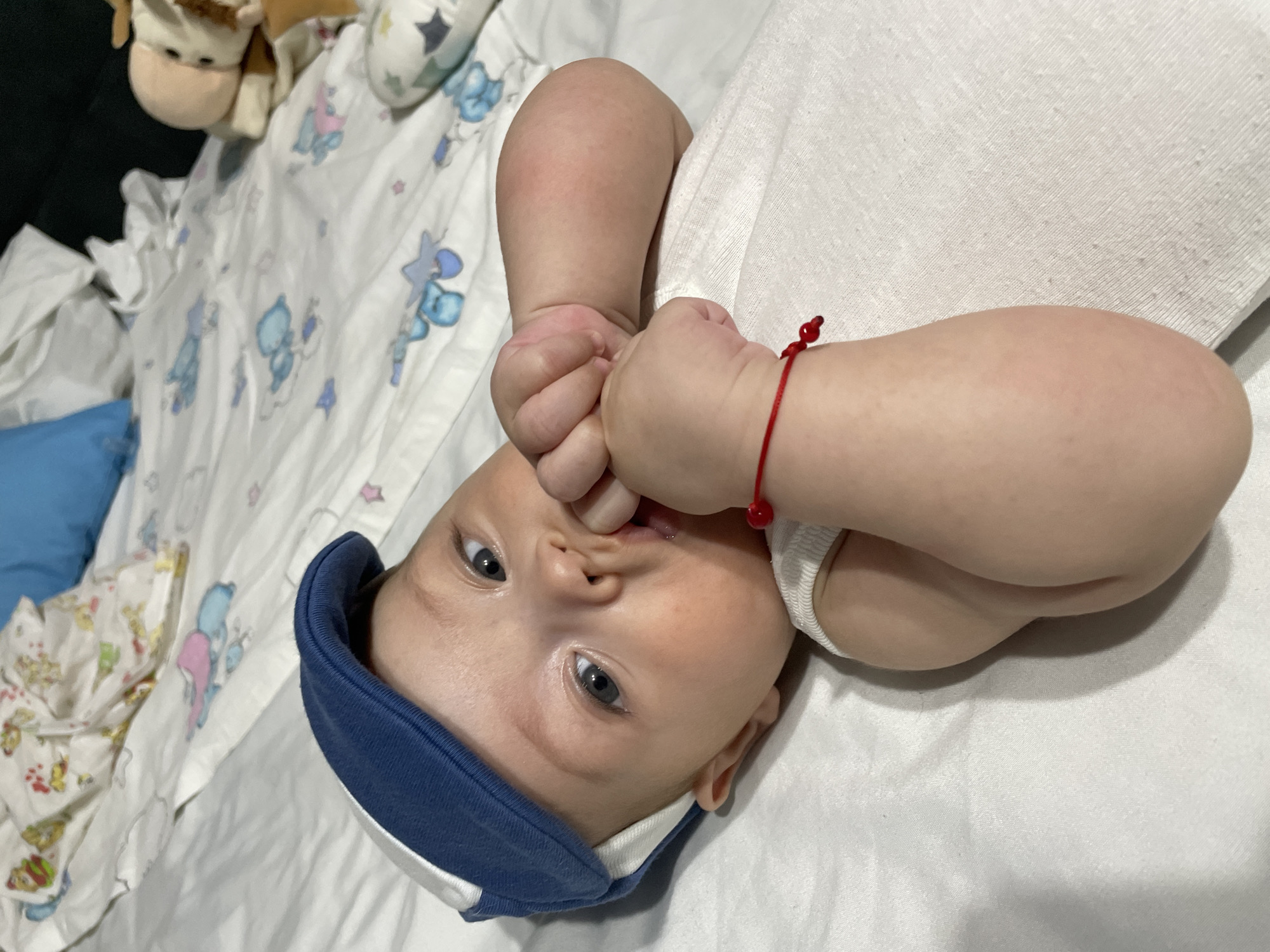 The image shows a close-up, overhead view of a baby lying on a white surface, possibly a bed or changing table, adorned with a blue and white patterned sheet featuring cartoon animals like bears and stars. The baby is wearing a white onesie and a blue and white soft hat, tilted slightly to the side. Their hands are brought up to their mouth, with fingers in their mouth, suggesting they might be teething or exploring their hands. The baby has light skin, bright blue eyes, and light brown hair. A thin red string bracelet with a small red bead is visible on their right wrist. In the background, partially out of focus, are some stuffed animals and a blue pillow. The lighting suggests an indoor setting, likely during the day, with no discernible weather conditions. The overall mood is tender and intimate, capturing a moment of infancy. There is no visible text in the image. The location context is Fălești, Moldova.