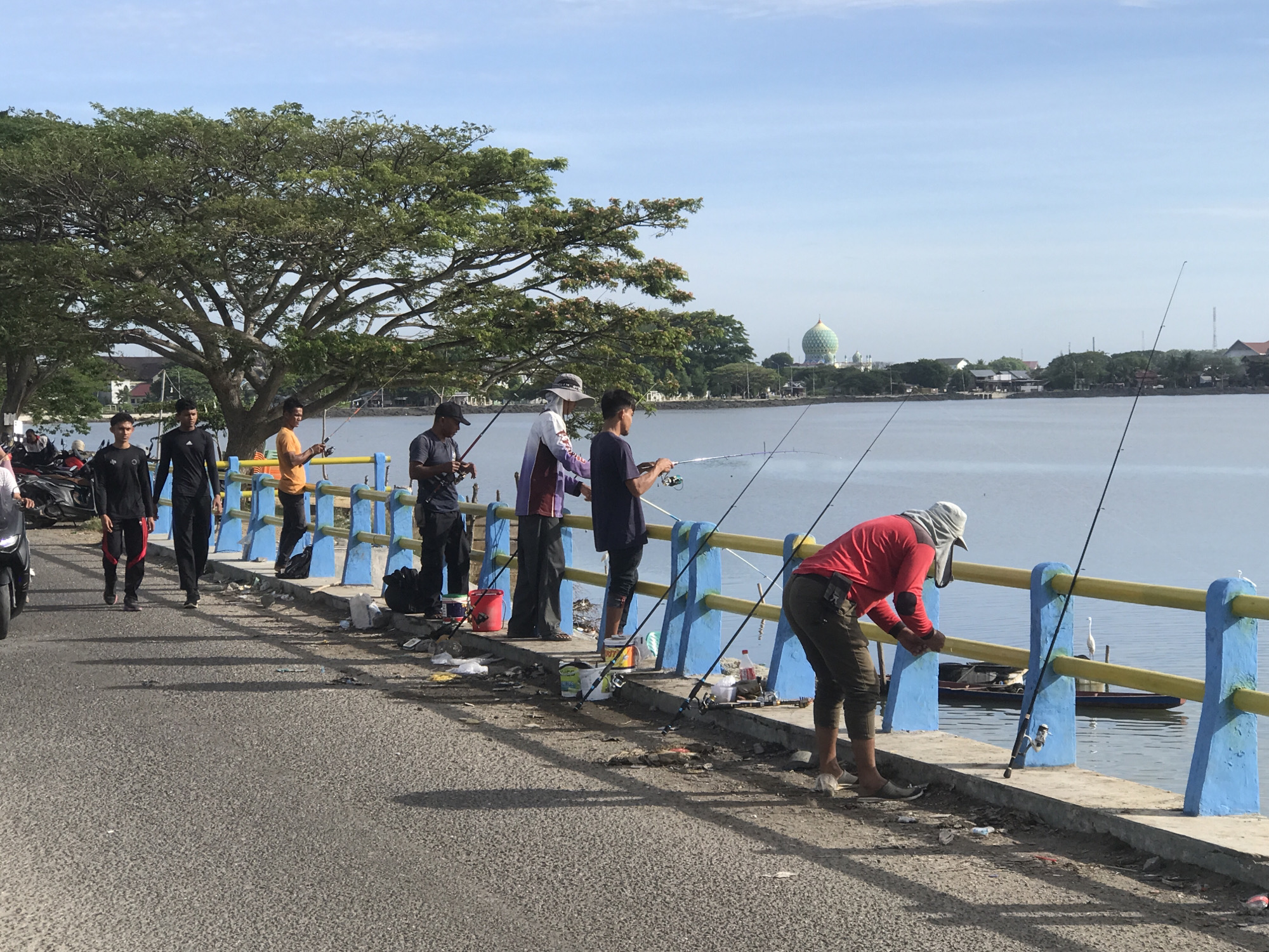 The scene captures a group of men fishing from a bridge in Lhokseumawe, Indonesia. Seven men are visible: two are walking along the road, while the other five are actively engaged in fishing over the blue and yellow railing of the bridge. Each fisherman has a fishing rod extended over the water. Fishing equipment such as buckets and tackle boxes sit nearby. Litter is scattered along the edge of the bridge.
In the background, a large tree extends its branches over the scene. Across the water, buildings and a distinct dome-shaped mosque are visible against a clear sky. The day appears bright. The road leading onto the bridge is gray asphalt. A motorcycle is parked on the side.