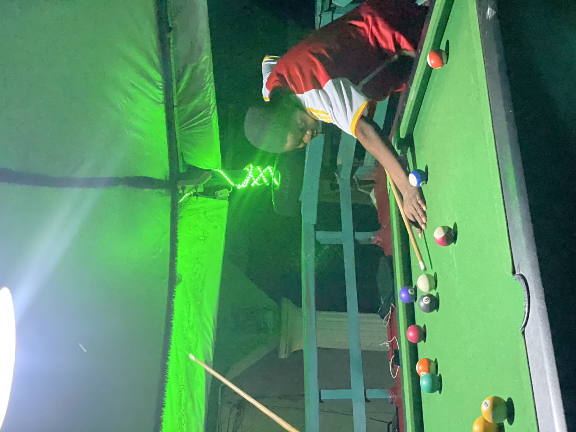 This image captures a person playing billiards on a pool table within an enclosed, dimly lit space in Lokoja, Nigeria. The scene is dominated by a strong green ambient light originating from multiple sources, casting a verdant hue across the setting.
One individual, appearing to be a young person with dark skin, is positioned on the right side of a standard-sized pool table with a green felt surface and dark rails. The person is wearing a short-sleeved shirt, predominantly red with white shoulders/sleeves and yellow stripes. They are bent over the table, holding a pool cue with both hands, poised to strike a white cue ball. The cue ball is located near a black 8-ball and a solid purple ball. Other billiard balls scattered on the table include solid blue, green, yellow, orange, and red balls, as well as striped red/white and orange/white balls. Another pool cue leans vertically against a wall or structure on the far left.
Overhead, a bright white light fixture is visible in the upper-left corner, providing primary illumination. The ceiling appears to be draped with light-colored fabric or tarp, which reflects the pervasive green light. In the background, green LED string lights are arranged in an "X" pattern on a support structure. A teal-colored railing or partition runs horizontally in the mid-ground. Faintly visible text "ZEALOT" appears on a dark object, possibly an electronic device, behind the player. Various other objects, including cables and what appears to be a bag, are on a surface behind the railing.