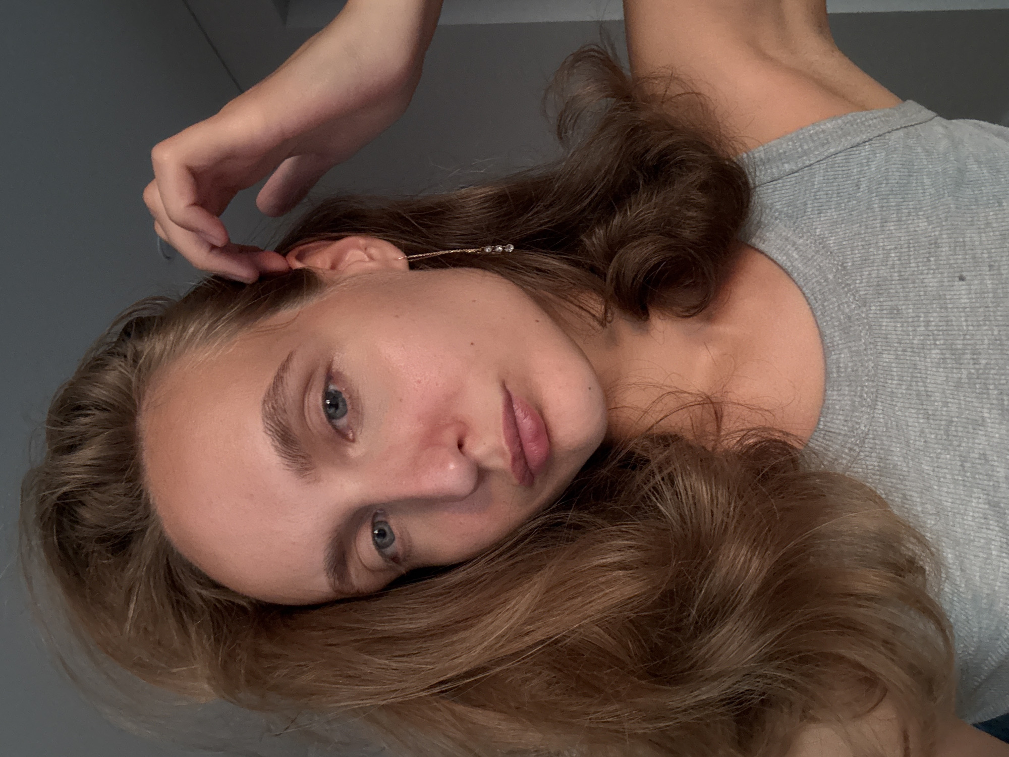 A young woman with long, wavy brown hair and blue eyes is pictured in a close-up shot. She is wearing a grey tank top and a delicate gold earring with three small clear stones. Her right hand is raised, touching her hair near her ear. Her skin appears fair and slightly flushed on her cheeks. The background is a plain grey wall. The image appears to be taken indoors. There is no indication of the location being in the city of Подшивалово, Russia.
