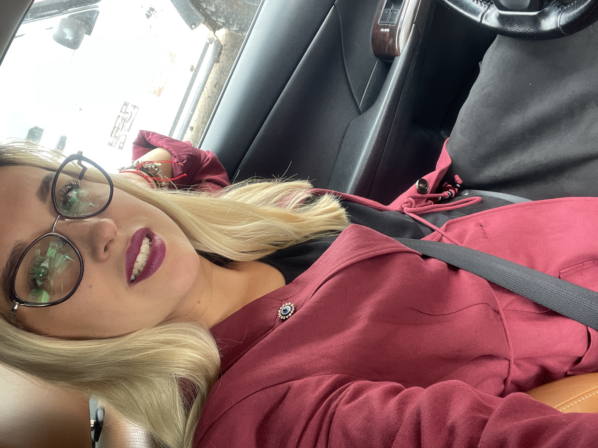A young woman with blonde hair and glasses is taking a selfie inside a car. She is wearing a maroon jacket with a blue and silver brooch. Her lips are painted a dark berry color, and she is smiling slightly. The interior of the car is visible, including the steering wheel, dashboard, and car door. The woman's bracelets are also visible on her wrist. The scene appears to be taking place during the daytime, and the weather is likely sunny, as indicated by the bright light reflecting on the windshield. The location is Chaikovsky, Russia.