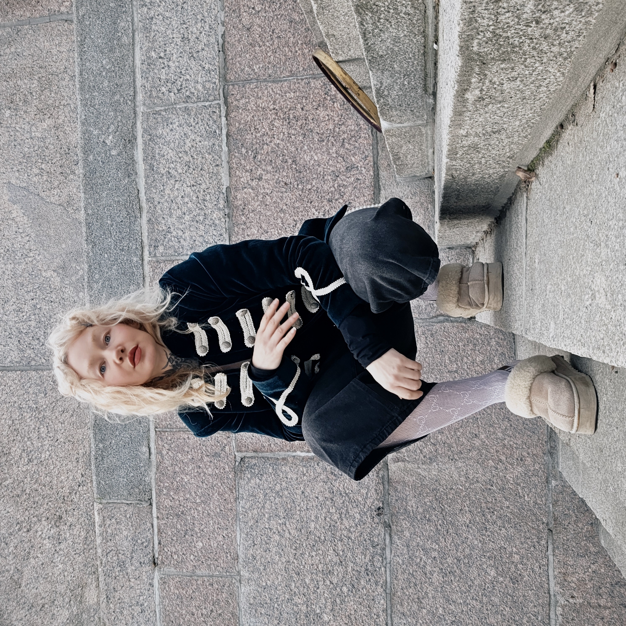 A blonde woman wearing a navy velvet military style coat with a white rope closure, dark pants, patterned tights, and tan sheepskin boots is crouching on granite stairs. The location is Saint Petersburg, Russia. A broken wooden frame leans against the stair wall in the background. The environment consists of granite steps and walls.