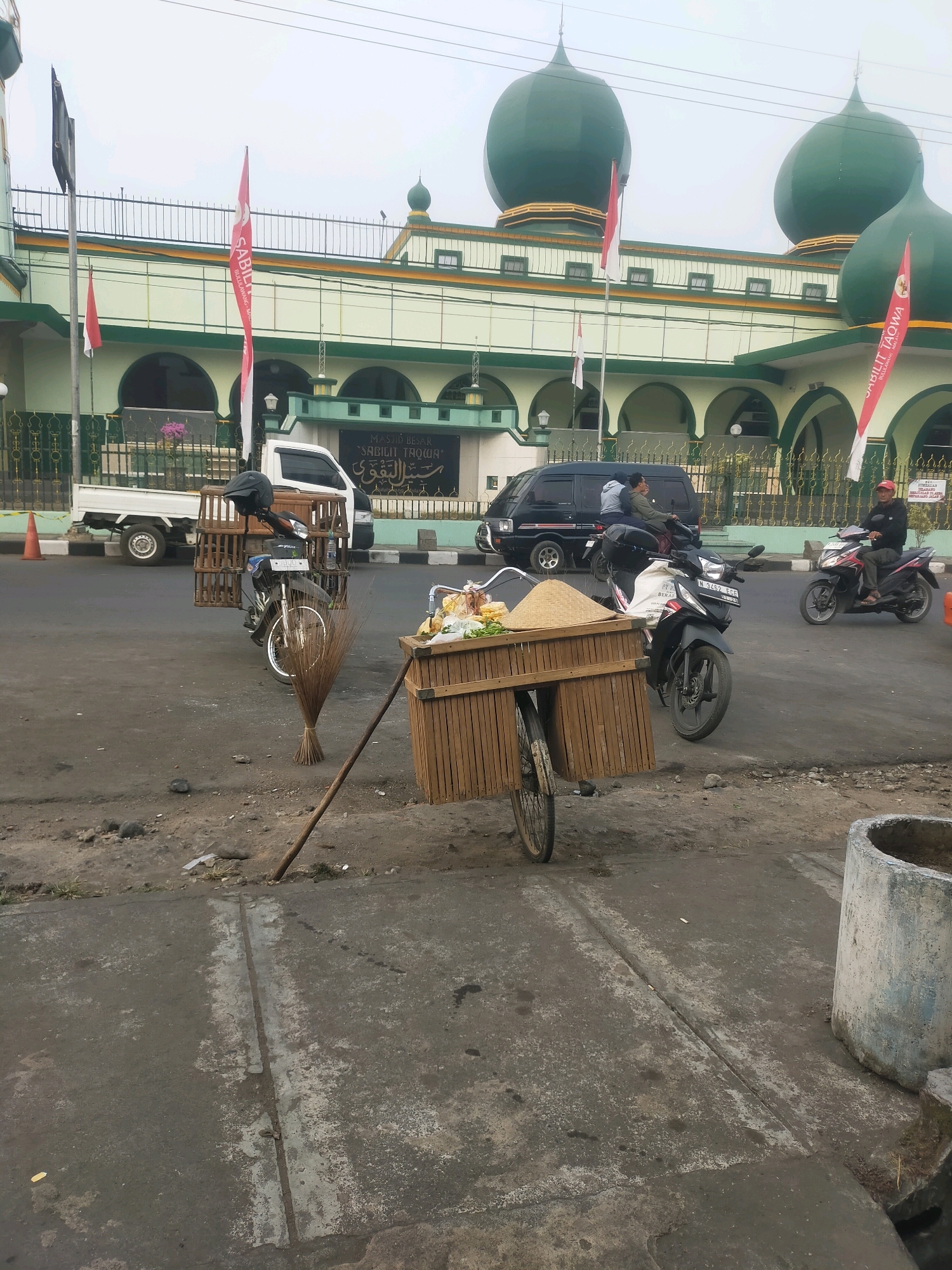 The image depicts a street scene in front of a mosque in Bululawang, Indonesia. The mosque, identified by the signage as "MASJID BESAR SABILIT TAQWA," has green domes and a light green facade with arched windows. In front of the mosque, a white pickup truck is parked.
In the foreground, a weathered wooden cart is positioned on the dirt and asphalt ground. The cart is laden with various items, including a conical woven hat, what appears to be produce like bananas and leafy vegetables, and a pile of grain or sand. A broom is leaning against the cart, with its bristles fanned out on the ground.
A motorcycle, also in the foreground, is parked near the cart and broom. It has a wooden cage structure attached to its rear, possibly for transporting goods or animals. A helmet rests on the motorcycle seat.
Several other vehicles and individuals are visible on the street. A black van is parked behind the white pickup truck, with two people riding on a motorcycle in front of it. Further down the street, another motorcycle is being ridden by a person wearing a red cap and a light-colored shirt. Indonesian flags, red and white, are prominently displayed on poles in front of the mosque. The overall environment suggests a public area with some commercial activity taking place.