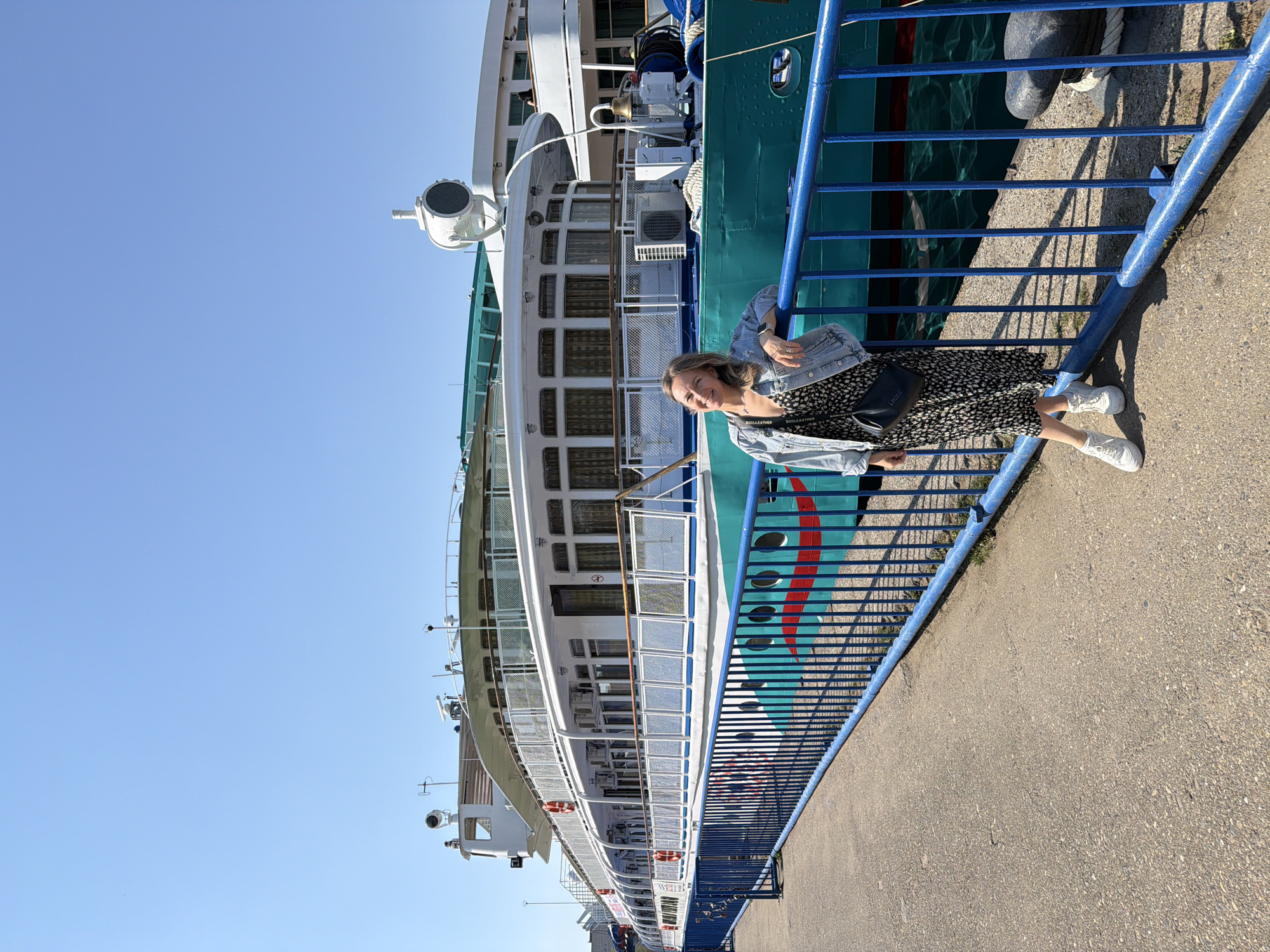 A medium shot captures a woman leaning against the blue railing of a large, multi-story ship docked at the waterfront in Chaikovsky, Russia. The ship is painted in a combination of white, turquoise, and green colors. The woman has blonde hair and wears a black and white patterned dress, a light blue denim jacket, and white sneakers. A black purse hangs on her shoulder. She is smiling towards the camera. The blue railing separates the ship from a concrete walkway. Above the ship, a clear blue sky is visible.