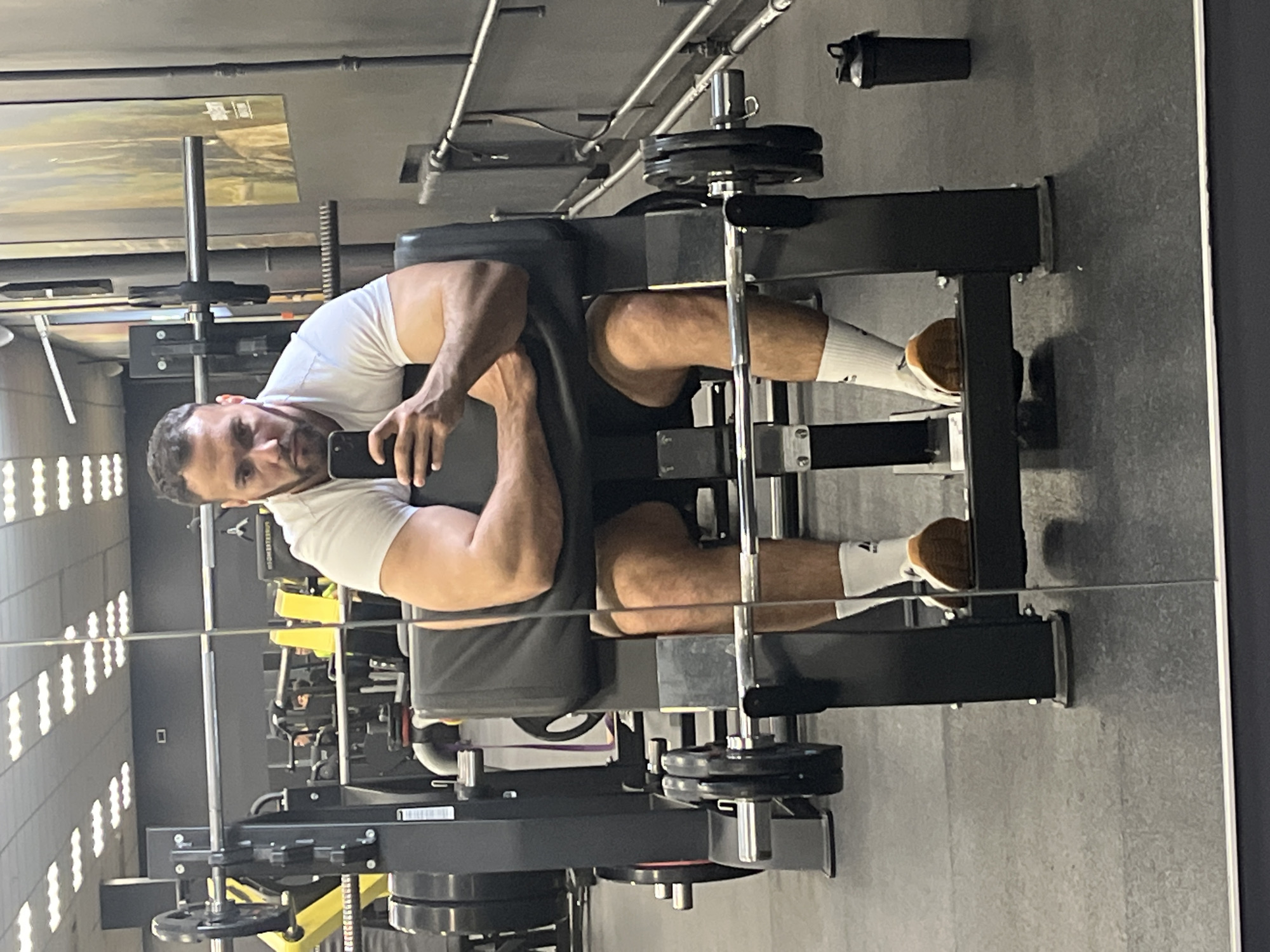 A man in a white t-shirt and dark shorts is seated on a leg press machine at what appears to be a gym. He is wearing white socks and light brown sneakers. He is holding a cell phone in his left hand, presumably taking a photo of himself in a mirror that reflects the scene. The leg press machine is loaded with weight plates. A black water bottle is visible in the upper right corner. The gym has windows and appears to be in a commercial building in Ulyanovsk, Russia.