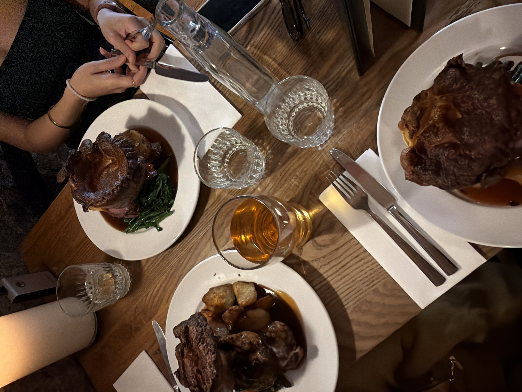 A high-angle shot captures a dinner table setting, likely in Unknown, Unknown, featuring three plates of food. Each plate contains a generous portion of roast meat, Yorkshire pudding, and vegetables, possibly spinach or kale. A glass of amber liquid, possibly beer or wine, sits near one plate, alongside a water carafe and a smaller glass. Cutlery is neatly arranged on napkins beside each plate. Two individuals are visible; one is holding silverware and preparing to eat, while another’s arm enters the frame from the bottom right. The overall ambiance suggests a meal being shared in a restaurant or dining establishment.