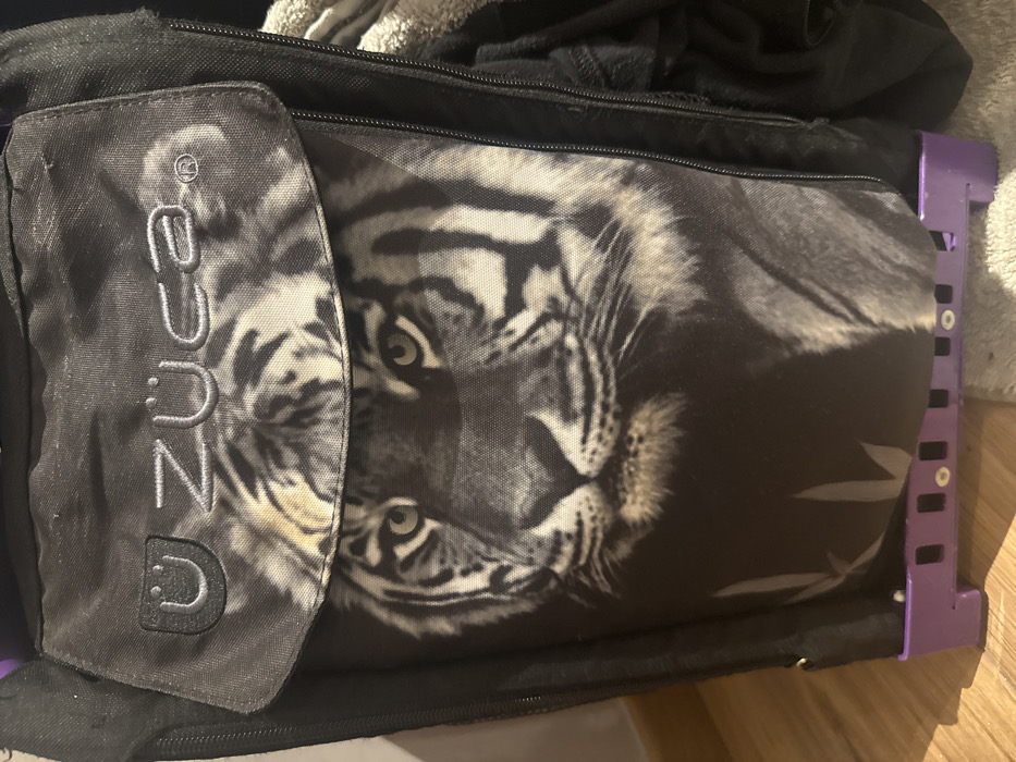 The image shows a Zuca brand rolling bag with a tiger print design. The tiger's face is prominently displayed on the main body of the bag. There is a front pocket with the Zuca logo vertically embroidered. The bag has a black trim, a purple metal frame, and is sitting on what appears to be a wooden floor. Clothes or other items are visible on top of the bag.