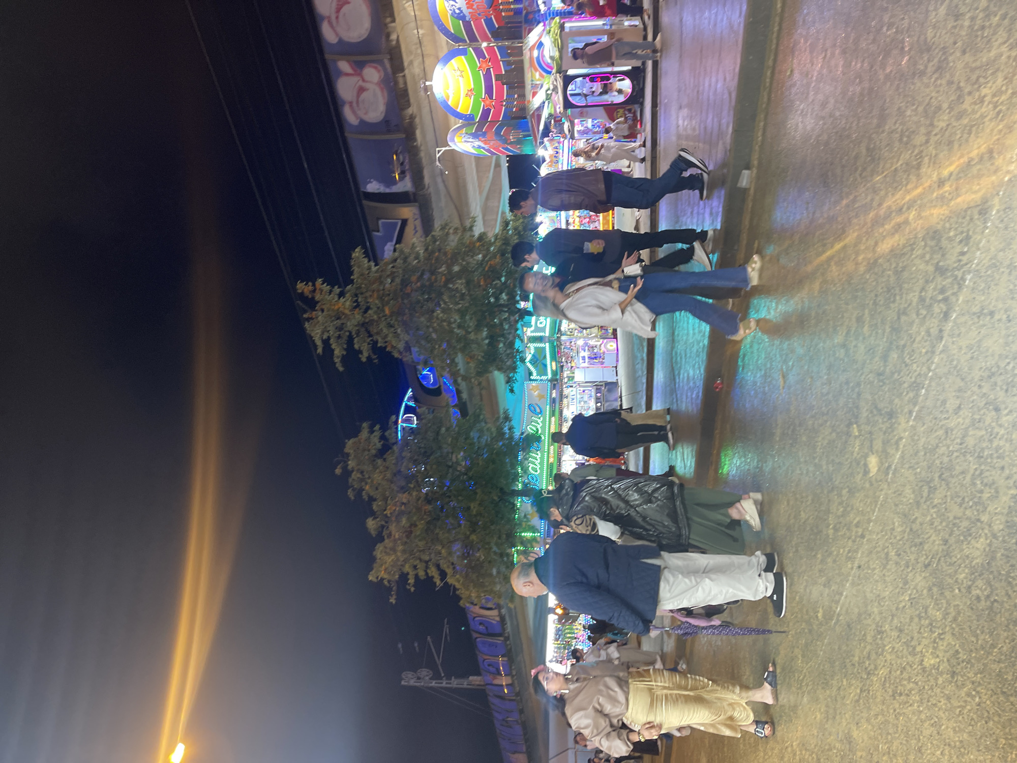 At an amusement park in Jerez De La Frontera, Spain, people are milling about in the early evening. Individuals are strolling, some in groups, on a paved surface. Trees are interspersed among the amusement park attractions. The night sky is visible. A beam of light shoots upward.