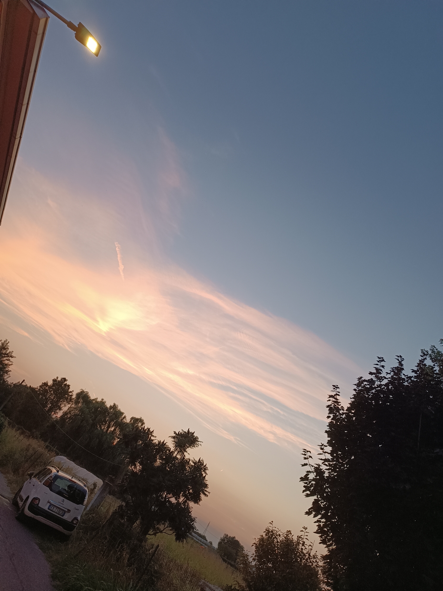 This image depicts a sunset scene in Viadana, Italy. The sky is a gradient of blue and pink, with wispy clouds. A street lamp is illuminated in the upper left corner. In the foreground, a white car is parked on a dirt road, with tall grass and trees surrounding it. The car appears to be a compact hatchback, possibly a Citroën C3 Picasso, with the license plate visible. The overall atmosphere is serene and rural, suggesting the outskirts of the city. There are no people visible in the image.
