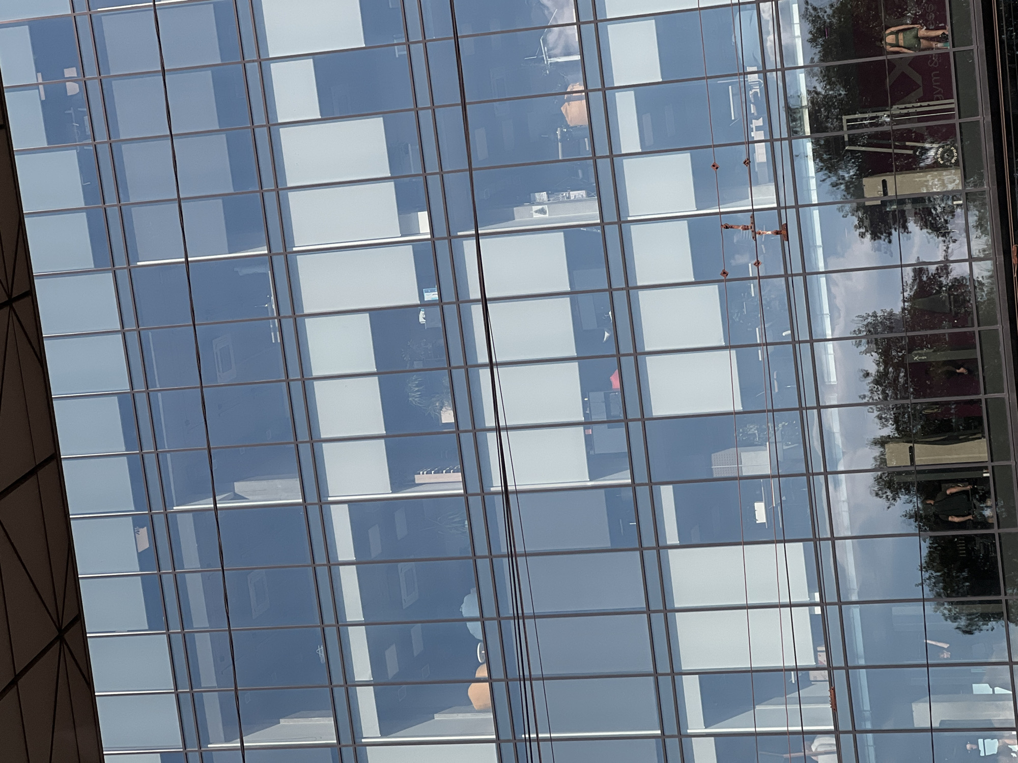 This media file shows a close-up, rotated view of a modern glass-facade building. The building appears to be an office building or a commercial complex with multiple floors. The glass panels reflect the sky, trees, and possibly other buildings, creating a mosaic of light and shadow.
Within the reflections, there are glimpses of interior spaces, including office desks, chairs, and what appear to be potted plants. The reflections are fragmented and distorted, making it difficult to discern specific details of the interior activities. However, some reflections suggest the presence of people inside the building, though their activities are unclear.
In one section of the reflection, a person in athletic wear, possibly a woman in shorts and a tank top, is visible in what could be a gym or fitness center, given the presence of gym equipment like benches and weights. Text is partially visible within this reflection, including "Gym & Fitness."
The lighting suggests it is daytime, with the sun casting bright reflections on the glass. The weather appears to be clear and sunny, indicated by the bright reflections and blue sky. The overall scene conveys a sense of urban business or activity.
While the location context is Ternopil, Ukraine, there are no explicit visual cues within the image that definitively confirm this specific location. The building's architecture is generic to modern urban design.