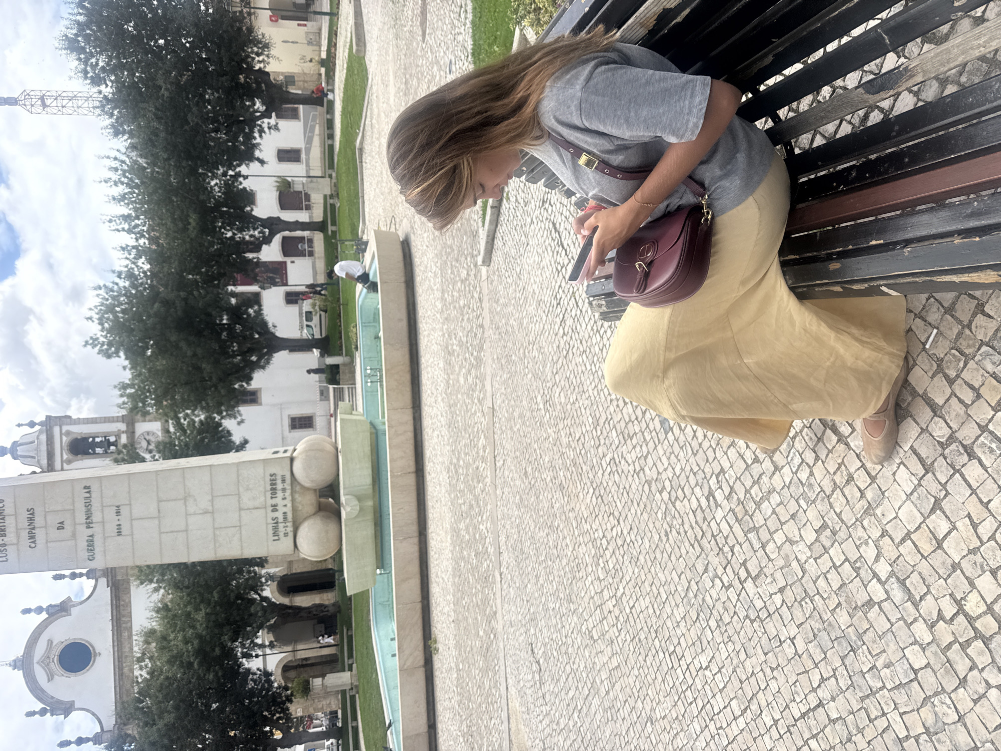 A young woman with long brown hair is sitting on a dark wooden park bench in a city square. She is wearing a grey t-shirt and a long, flowing yellow skirt. A burgundy shoulder bag rests on her lap. She is looking down at her mobile phone, which she is holding in her hands. The bench is positioned on a cobblestone pathway, with a green grassy area and a light blue fountain in the background. Behind the fountain, a large white monument with inscriptions stands prominently. In the distance, there are trees and a white building with a bell tower, possibly a church. The sky is partly cloudy. The location is identified as the Praça do Município in Torres Vedras, Portugal, a significant historical site featuring the Monument to the Peninsular War and the former Town Hall building.