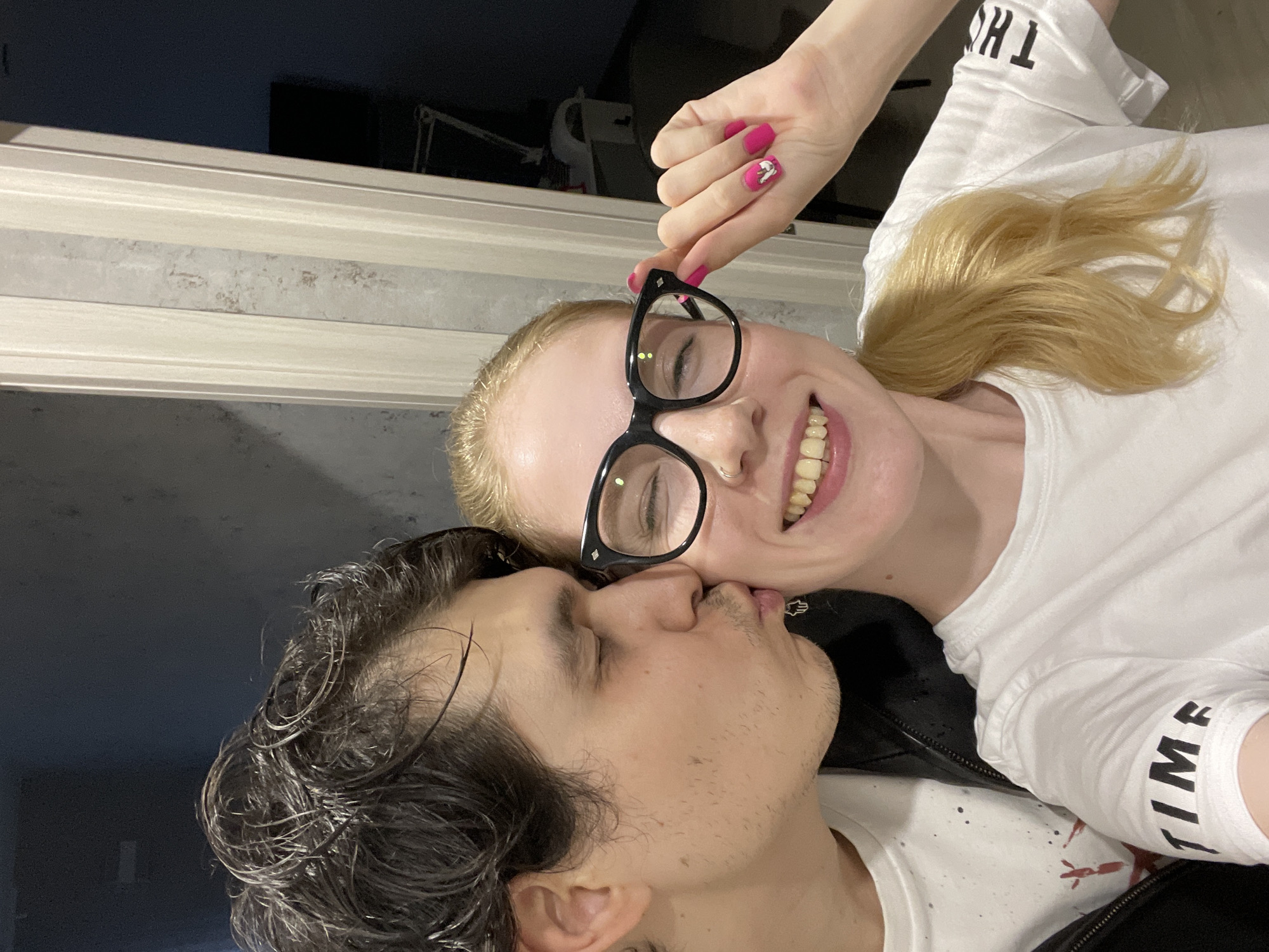A selfie depicts a woman with fair skin and blonde hair, wearing black-rimmed glasses and a white t-shirt, smiling and holding her glasses with a hand bearing pink nail polish. A man with dark, wet hair is kissing her cheek. The location cannot be determined from this image. No additional details can be extracted.