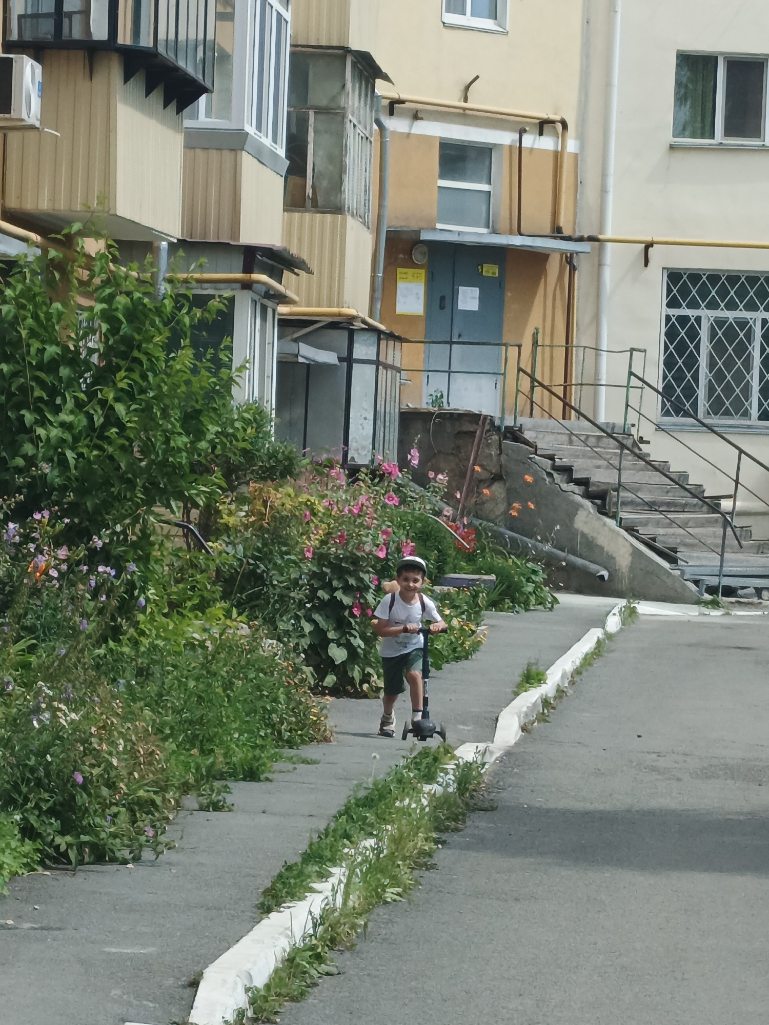 A young boy, wearing a white t-shirt, green shorts, and a white cap, rides a black scooter along a paved walkway next to a building. Lush green bushes and pink and purple flowers border the walkway on the left. To the right of the walkway, a concrete staircase ascends along the side of a building. The building has light-colored walls and several windows, some with white lattice patterns. An air conditioning unit is visible on the upper left portion of the building. The scene appears to be in a residential area in Kurgan, Russia, with the typical appearance of urban apartment blocks and vegetation common in Russian cities.