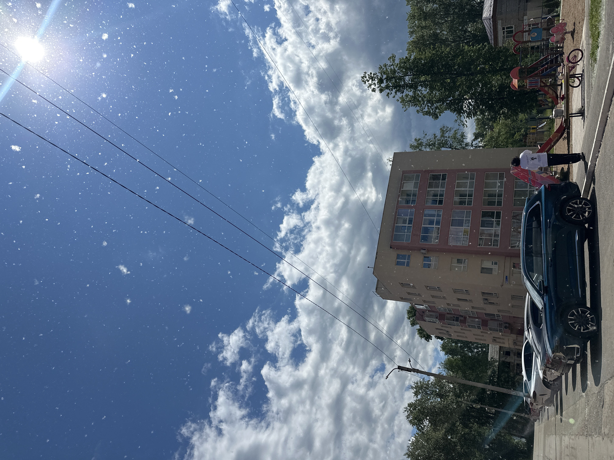 The image displays an outdoor urban scene in Chaikovsky, Russia, captured during daylight hours. The sky is largely blue with scattered, dense cumulus clouds and a prominent sun in the upper right quadrant, producing a strong glare and lens flare. Numerous small, white, blurry particles are airborne across the entire frame, suggesting poplar fluff, light snow, or similar suspended matter. Three black power lines traverse diagonally across the upper portion of the sky.
In the midground, a multi-story residential building with a light beige facade and reddish-pink sections is visible, featuring multiple windows and balconies. To the left of the building, a row of parked cars lines a paved area, with a dark green SUV in the foreground, followed by a silver sedan, a dark sedan, and a white SUV. A light-colored utility pole with a streetlight is positioned on the far left.
To the right of the main building, a children's playground occupies a dirt or sand area. Playground equipment includes a red and yellow slide structure and a blue and pink spring-rider. A small pink bicycle lies on its side next to a wooden bench, on which a light-colored object rests. A single individual, facing away from the camera, stands near the playground, wearing a white long-sleeved shirt with a dark design and dark pants. Green trees are visible on the periphery, behind the buildings, and adjacent to the playground. A smaller building with a reddish-brown tiled roof is partially visible behind the playground on the right.
