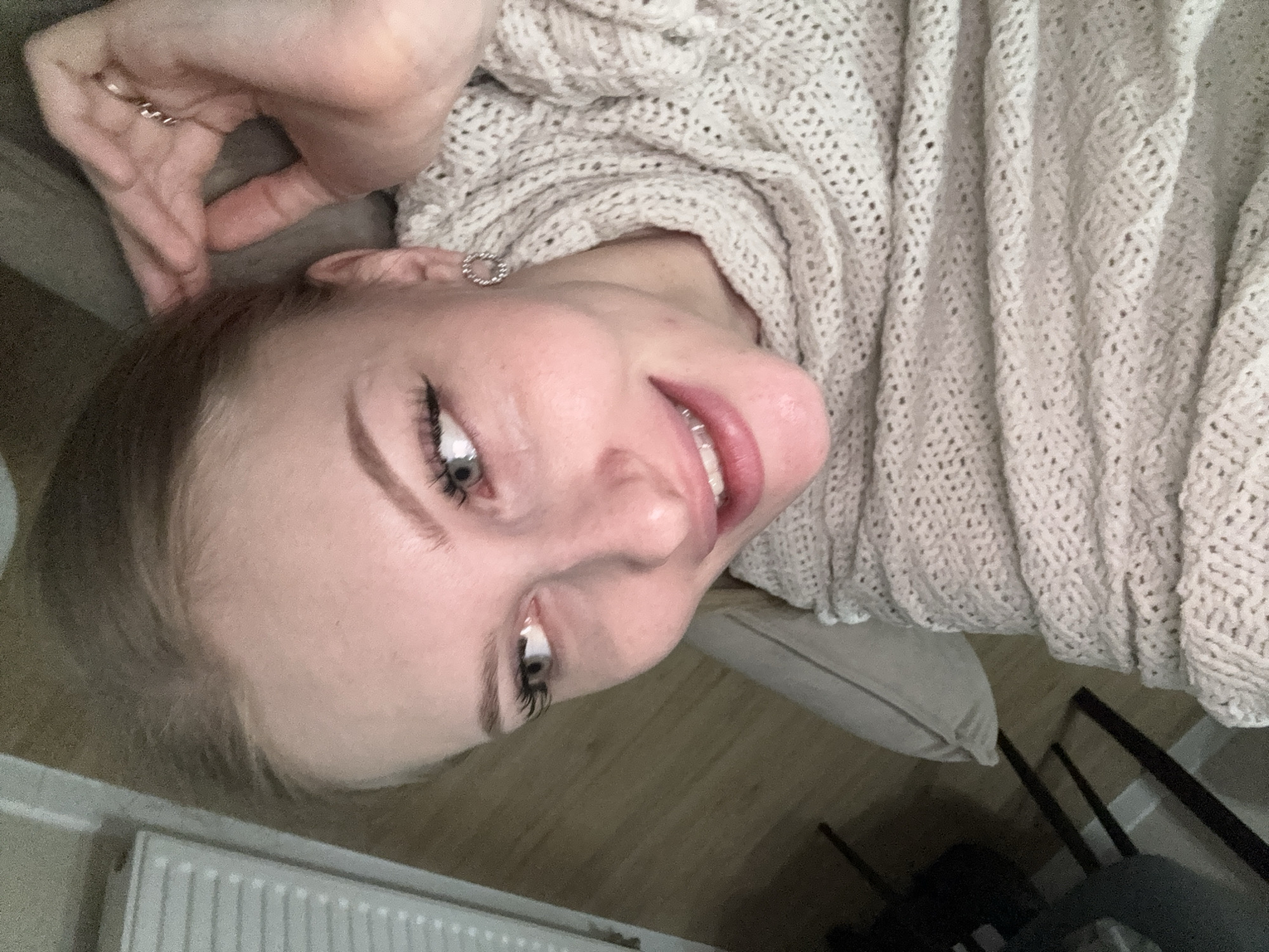 A woman with fair skin and blonde hair is lying down, smiling and looking up at the camera. She is wearing a beige knit sweater and a silver earring. Her hand is near her head, and she has a ring on her finger. The environment includes a radiator, a floor, a pillow and a chair. The location is Perm, Russia.