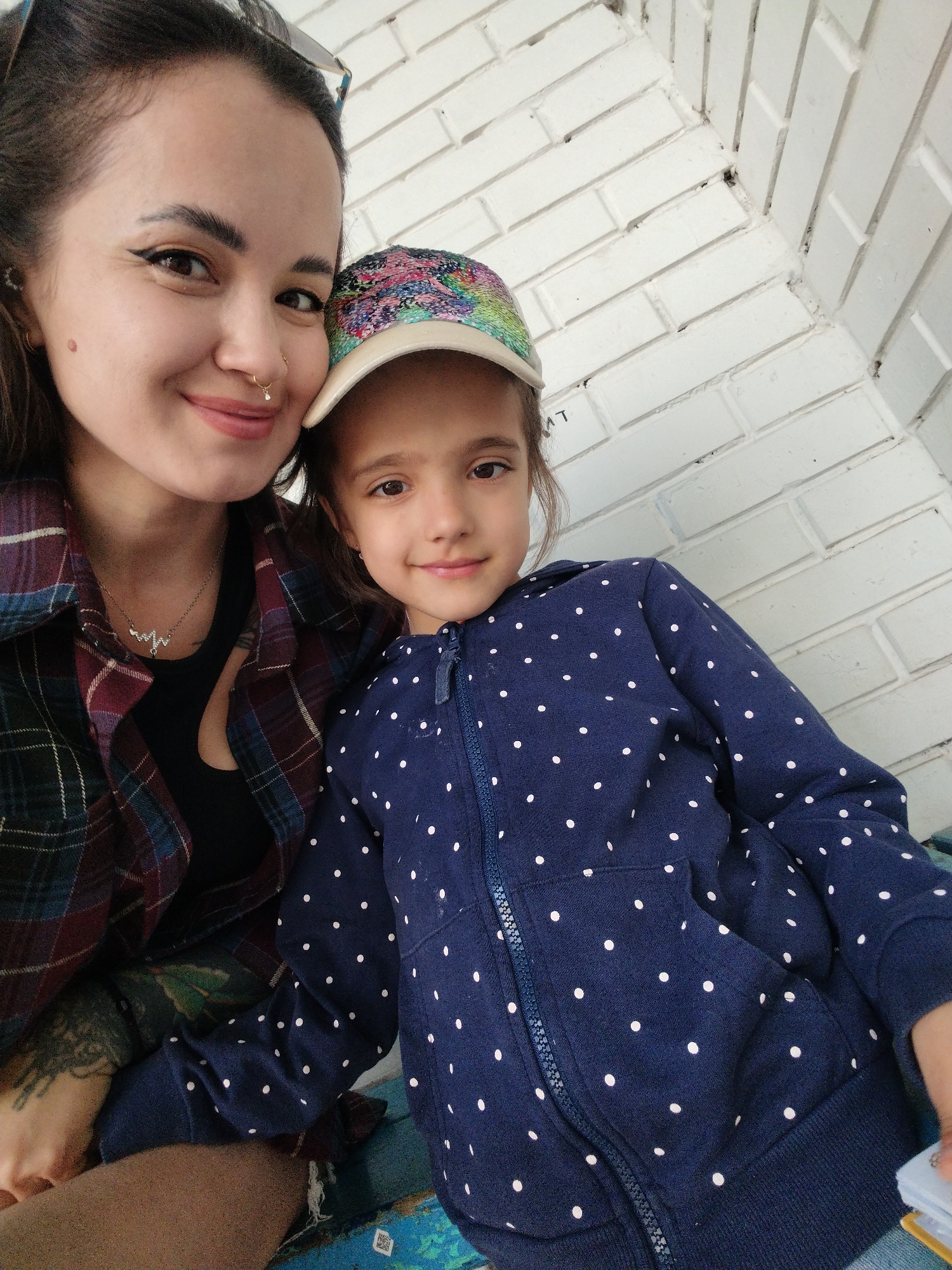 An adult female and a young female child are depicted in a close-up, selfie-style photograph. Both individuals are smiling with closed mouths and looking directly at the camera.
The adult female, positioned on the left, has dark hair, a prominent mole on her left cheek, dark winged eyeliner, and a small gold-colored nose ring with a clear stone on her left nostril. She wears a multi-colored plaid long-sleeve shirt in shades of maroon, dark blue, and light green, unbuttoned over a black undershirt. A silver-colored necklace with a stylized "M" or "W" pendant is visible against her black top. A dark-ink tattoo, resembling an organic or tree-like design, is visible on her left forearm.
The young female child, positioned on the right, wears a multi-colored, sequined baseball cap with a light-colored brim. She is dressed in a dark blue hooded jacket adorned with small white polka dots, featuring a central zipper closure and two front pockets. A small stack of rectangular objects, possibly cards or thin books, is visible in her right hand at the bottom right of the frame.
The background consists of a white-painted brick wall, which forms an internal corner behind the subjects. The bricks are laid in a stretcher bond pattern, and some minor discoloration is visible in the mortar lines and crevices. An inverted partial letter "T" in dark text is discernable on one of the white bricks behind the child's cap. The individuals appear to be seated on a textured surface, possibly a patterned textile like a blanket or cushion, featuring shades of blue and green with geometric motifs. A small white label, resembling a QR code, is visible on this surface in the bottom right. The scene is well-lit with soft, even light. This scene is located in the city of Noviy, Russia.