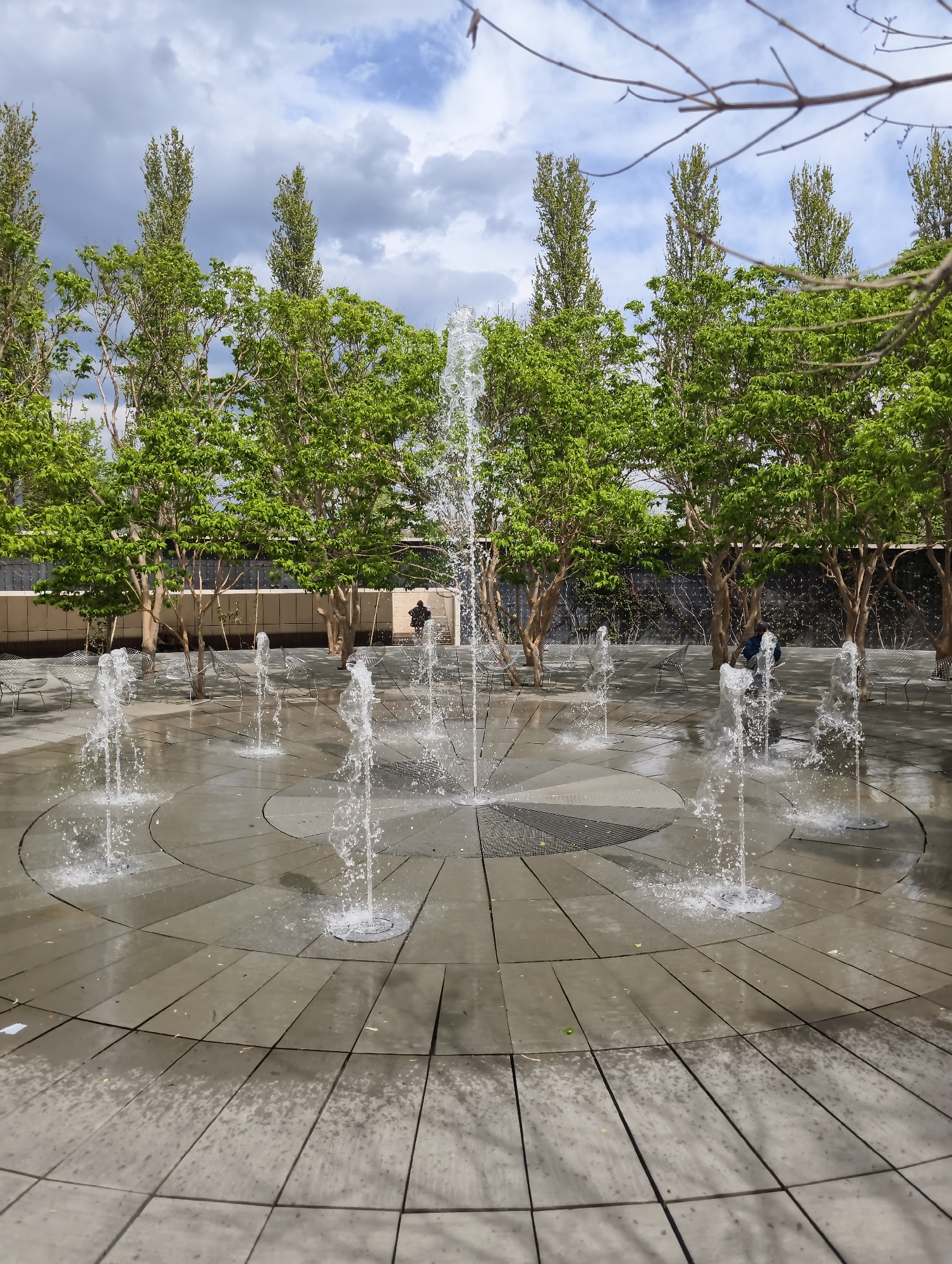 The image depicts a public space featuring a circular fountain with multiple water jets spraying upwards. The fountain is set within a paved area composed of concentric rings of rectangular slabs. Behind the fountain, a row of trees with green foliage lines the perimeter of the space. Further back, a wall or fence is visible. A few people are situated in the background, sitting on chairs or walking nearby. The sky is overcast with a mix of dark and light clouds. The scene is located in Krasnodar, Russia.