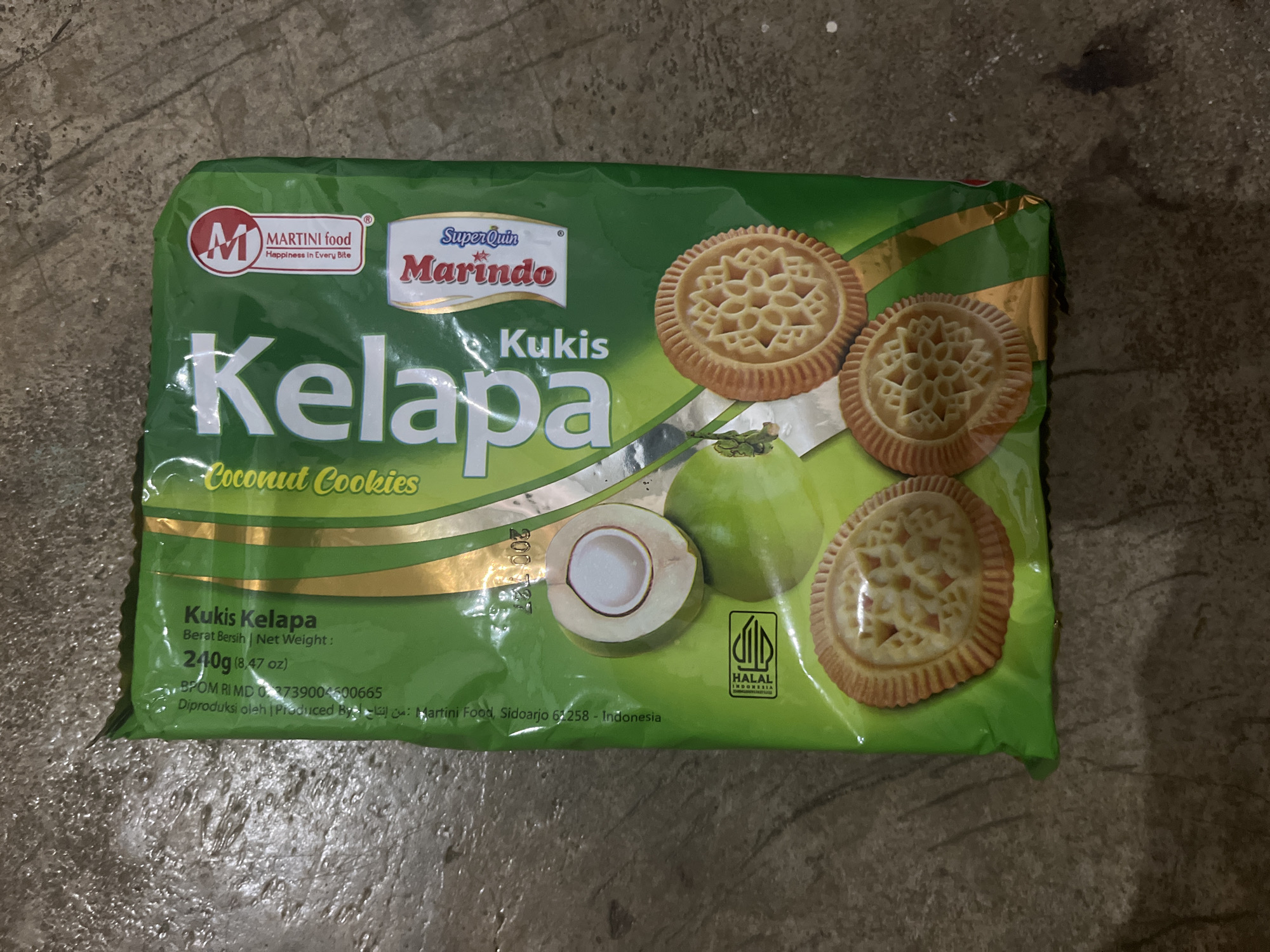 The image shows a green package of "Kelapa Kukis" (Coconut Cookies) made by "Marindo" and distributed by "Martini Food." The packaging features an illustration of coconut halves and several cookies. There is also a HALAL certification logo on the bottom left. The package appears to be lying on a textured, concrete-like surface. The location specified, Meliau Hulu, Indonesia, is not discernible from the visual information in the image.
