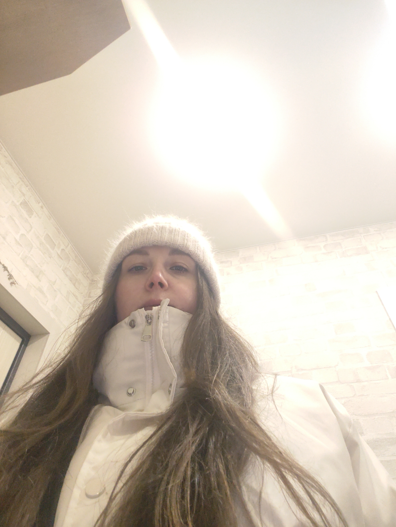 A selfie of a woman wearing a white, fluffy hat and a white winter coat with the zipper partially zipped. She has long brown hair. The background is a white brick wall and a white ceiling with an overhead light. The location is Unknown, Unknown.