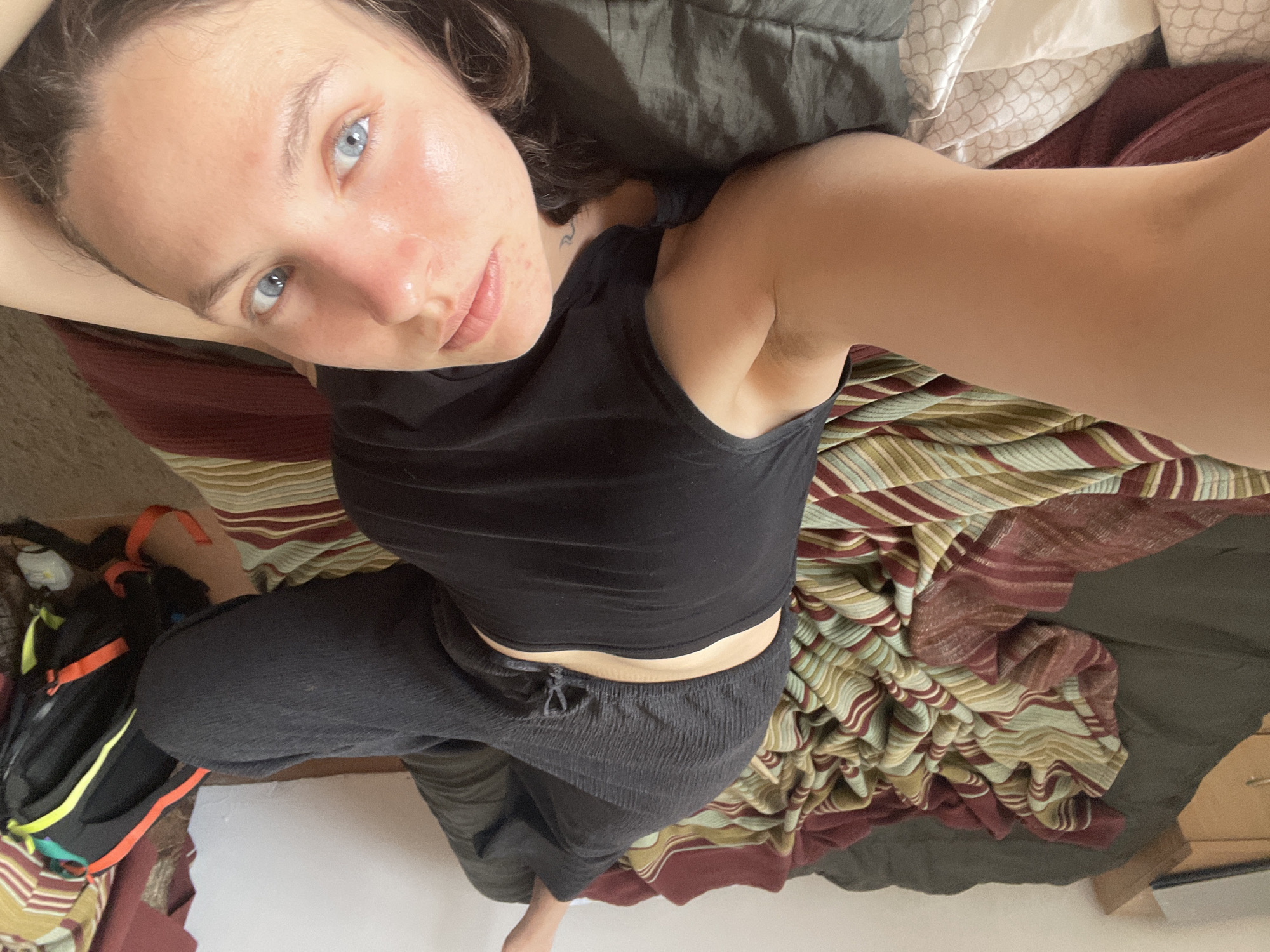 A young adult woman is lying on a bed with striped and maroon patterned sheets, as well as grey bedsheets. She is wearing a black tank top and dark-colored pants. A backpack is near her left foot. The location is uncertain, and there is no information to confirm that the location is Budva, Montenegro.
