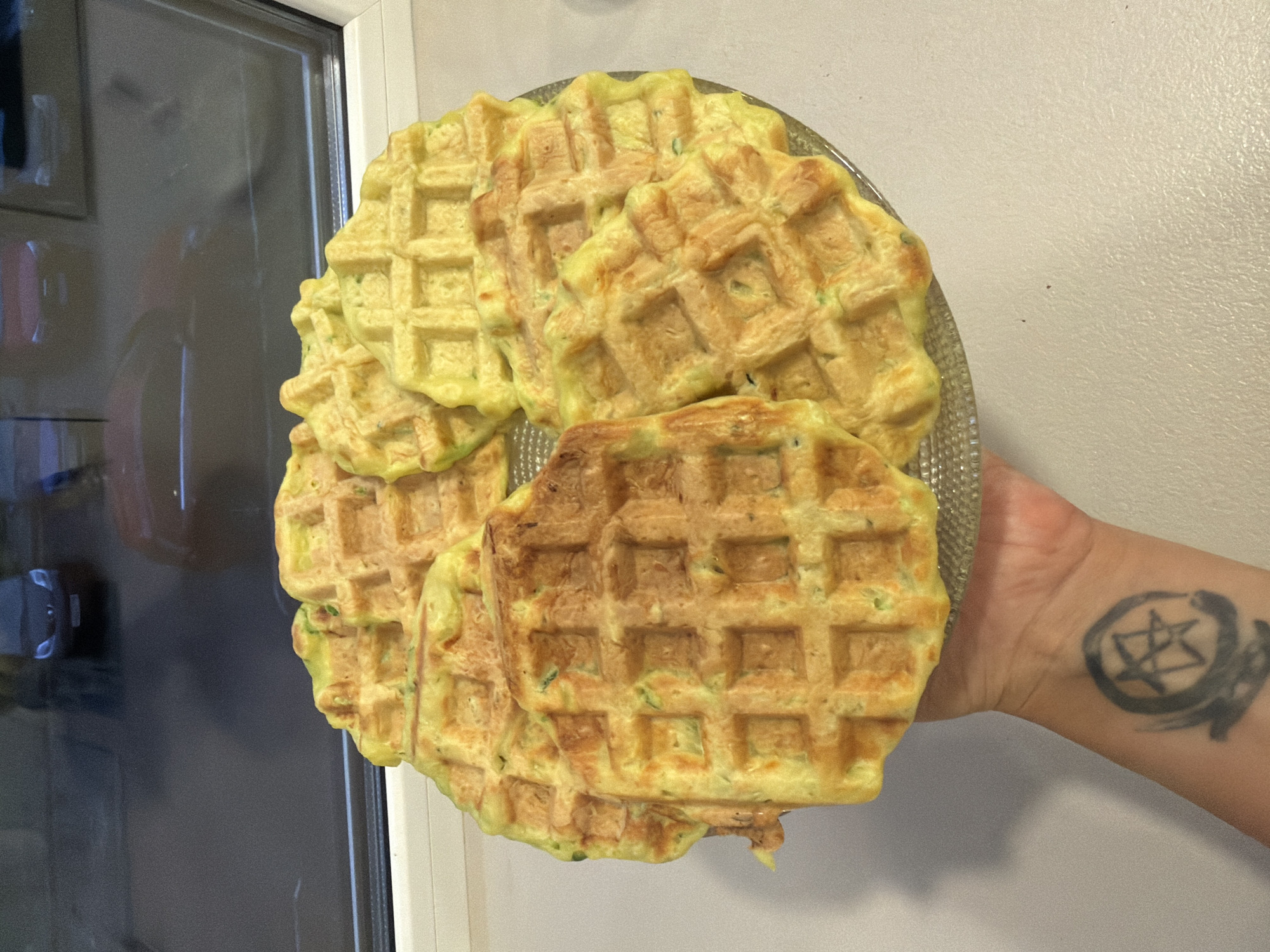 A plate of zucchini waffles is being held up in front of a window, reflecting cars in a parking lot. The waffles are golden brown with visible green flecks of zucchini. A person's tattooed arm is visible holding the plate. The scene is set indoors with a window overlooking an urban environment, possibly in Nefteyugansk, Russia.