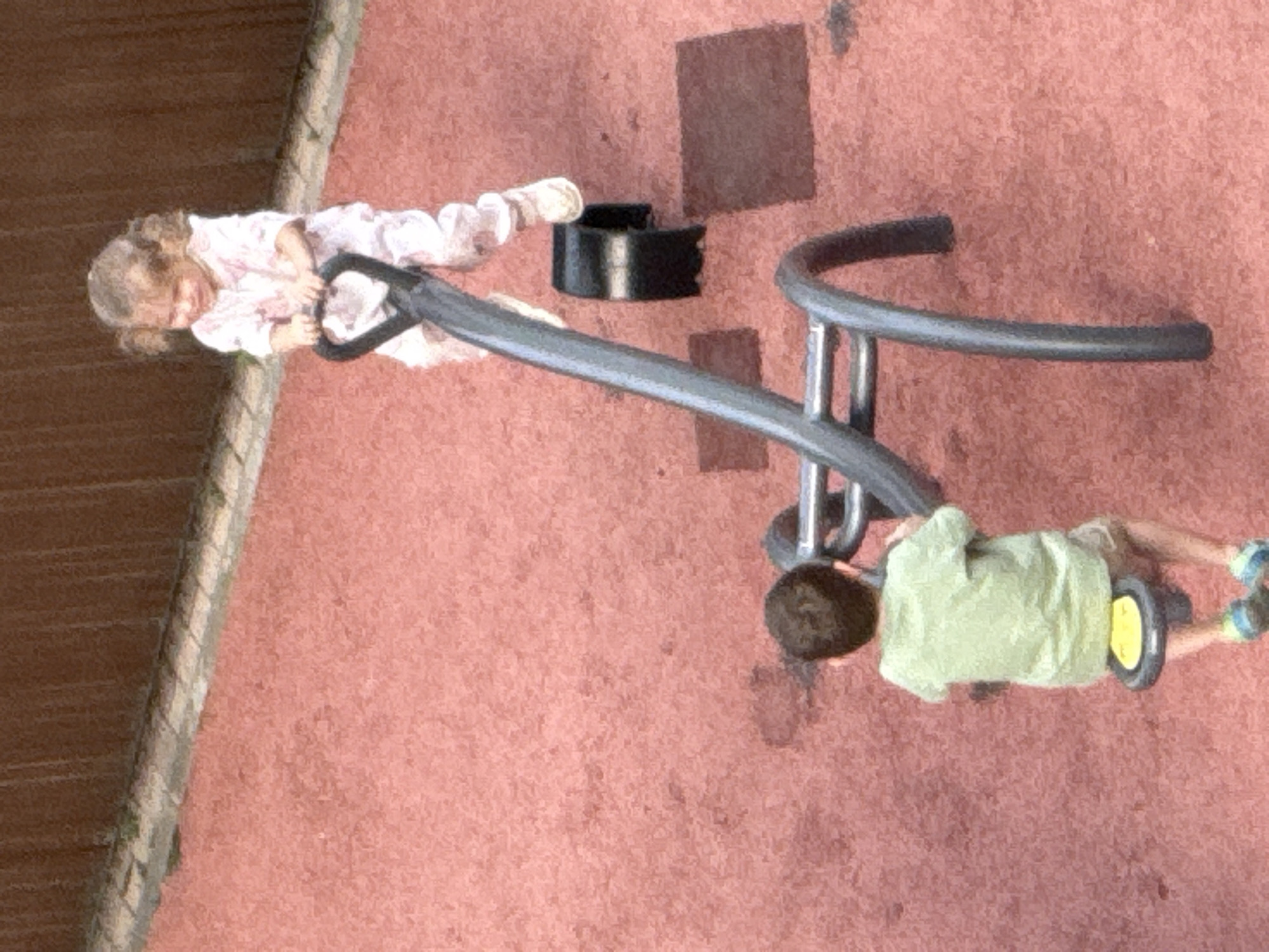 Two children are on a seesaw at a playground. A girl in a pink outfit is on the higher end of the seesaw, holding onto the handles. A boy in a green t-shirt and shorts is on the lower end of the seesaw, also holding onto the handles. The playground has a red rubberized surface and a wooden fence in the background. The image appears to be taken from an elevated perspective. The location is specified as Lobnya, Russia.