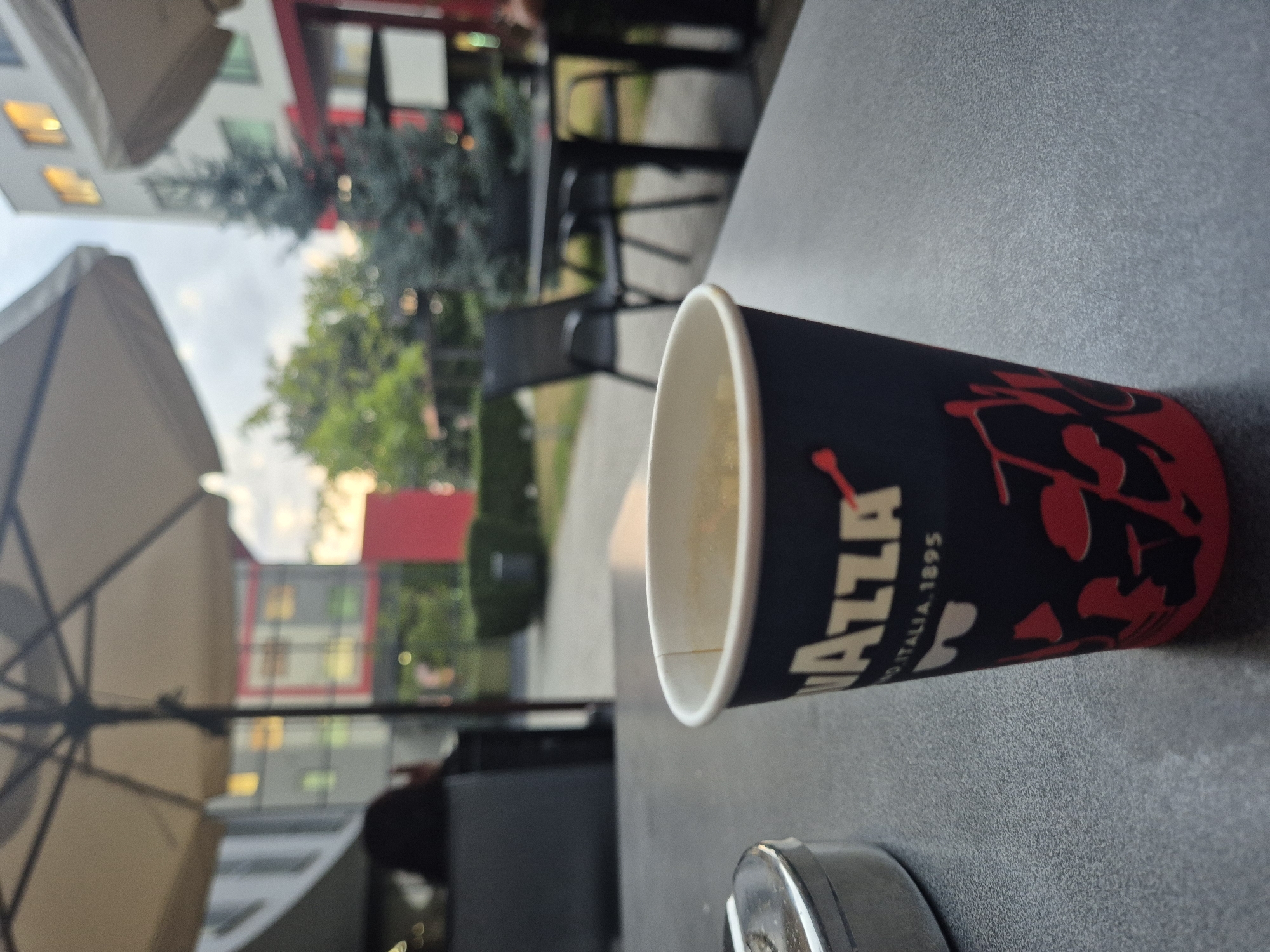 An outdoor seating area at a café features a close-up of a paper cup of coffee in the foreground. The cup is dark blue with a white "Lavazza" logo and red designs. To the left, a metallic ashtray is partially visible. In the background, tables and chairs are set up under umbrellas, with trees and modern buildings in the distance. The sky is overcast with hints of sunset. No people are clearly identifiable in the scene.