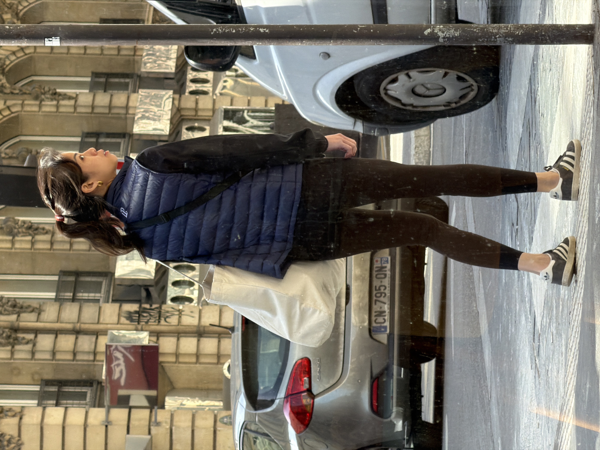 A young woman is reflected in a glass window in Paris, France, possibly a shop window or building façade, as she walks down a city street. She has dark hair pulled back into a ponytail with a red scrunchie, wears a blue puffer vest over a black top, black leggings, and black Adidas sneakers with white stripes. She carries a white bag over her shoulder. In the reflection, part of a white car is visible above her, and a silver car is behind her. The license plate of the silver car reads "CN-795-ON" with a French registration mark. Behind her reflection, the architecture of a Parisian building is seen, featuring multiple windows and ornate stone detailing on the facade. A sign with the word "Keras" is also visible on the building behind her reflection. The street itself and the cars on it suggest a typical urban environment.