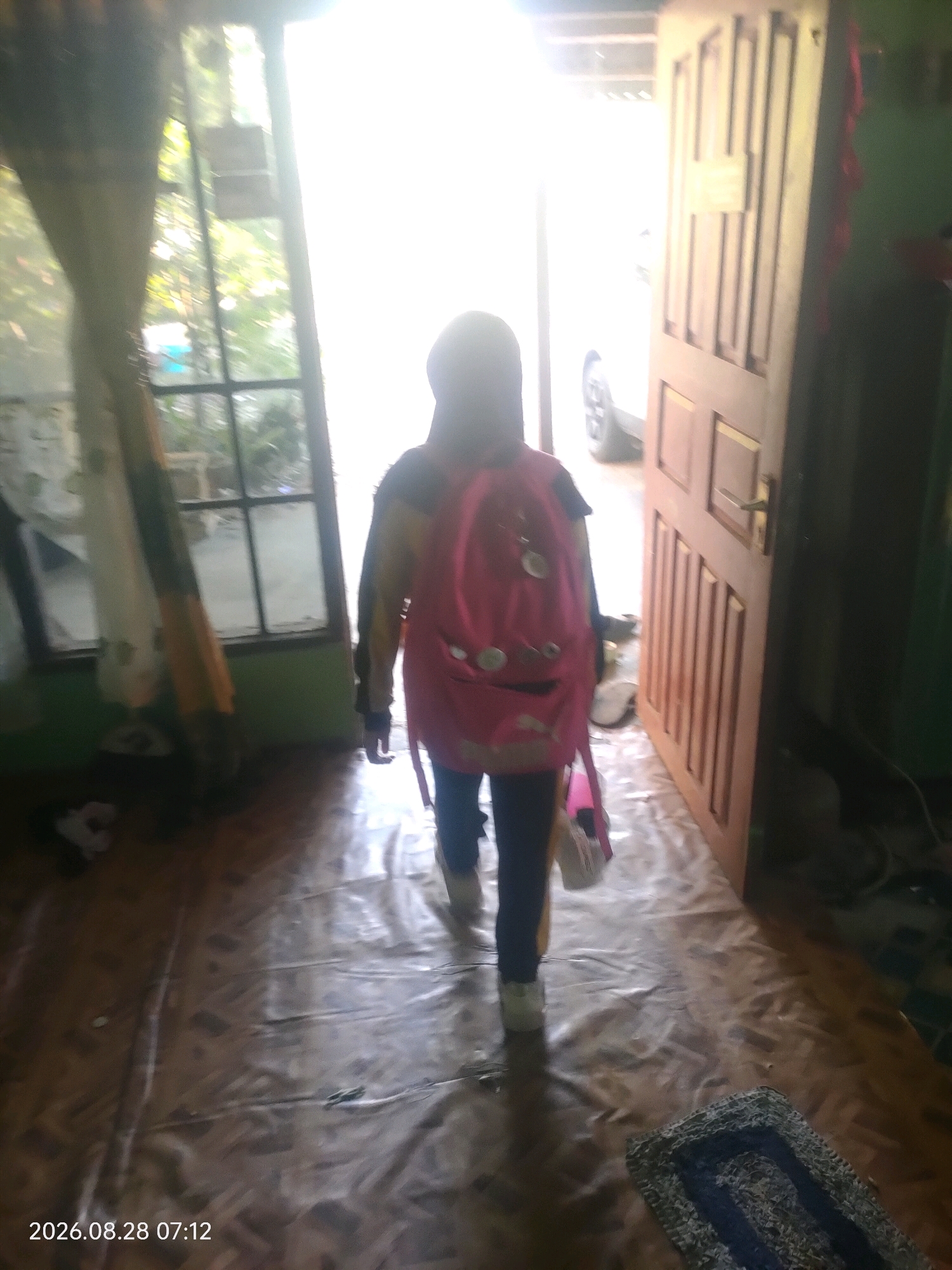 The image depicts the interior of a home in Kuaro, Indonesia, as indicated by the timestamp "2026.08.28 07:12". A young person, wearing a hijab and a backpack, is walking away from the camera and towards an open door. The backpack is a bright pink and appears to have some decorations on it. The person is wearing dark leggings and light-colored shoes. The environment is dimly lit, with natural light streaming in from the open doorway. The flooring is a patterned linoleum or vinyl. To the left, there is a window with curtains, and to the right, a wooden door is ajar. A car is partially visible through the open doorway, suggesting an outdoor area beyond. A small rug is visible on the floor in the foreground. The overall impression is a snapshot of a person leaving their home, possibly for school or an outing.