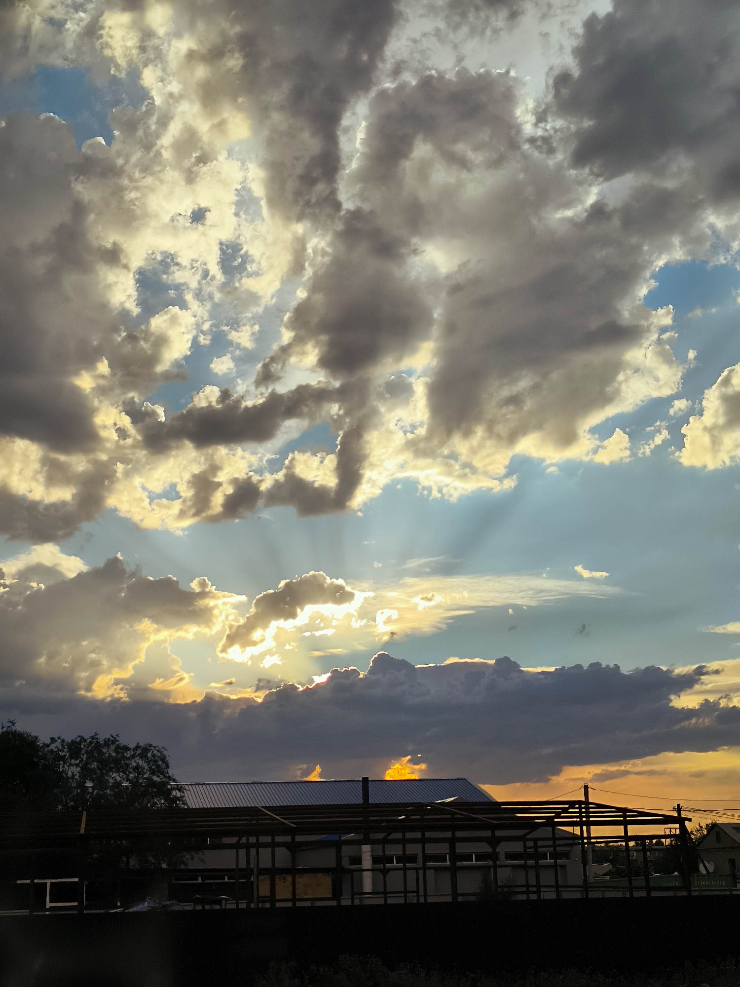 The image depicts a dramatic sky with heavy, dark clouds illuminated by shafts of sunlight, suggesting a sunset or sunrise. The clouds are varied in color, ranging from deep grays to golden yellows, and are broken by patches of blue sky. Below the sky, the silhouette of buildings and trees are visible. The foreground shows a dark, indistinct area, possibly vegetation or a fence. The architecture of the buildings appears industrial or under construction, with exposed frameworks and a metal roof. The overall atmosphere is one of natural grandeur, with the dynamic sky dominating the scene. The image does not provide specific details to confirm the location as Qarağandı city administration, Kazakhstan.