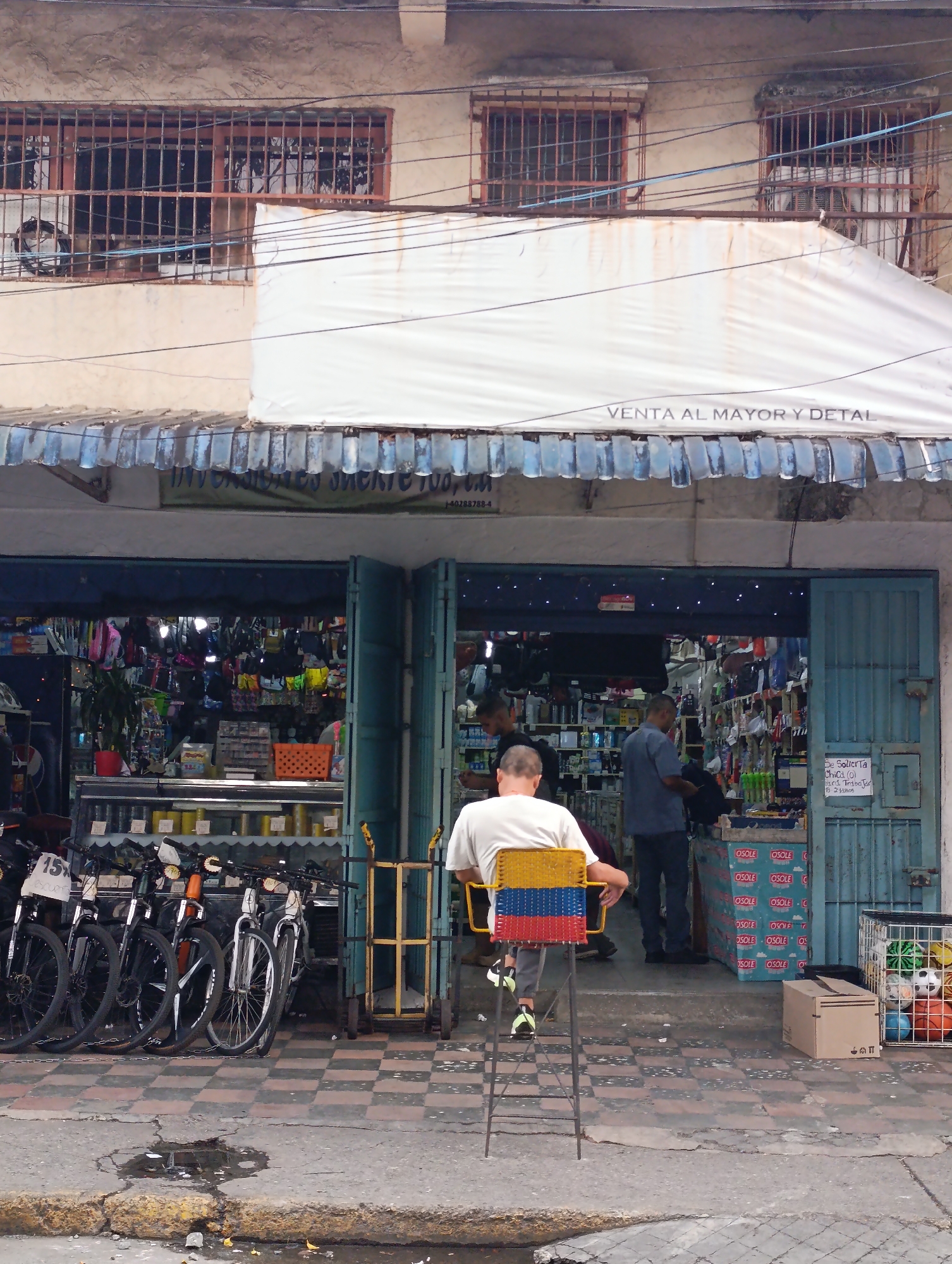 The image depicts a street scene in Tinaquillo, Venezuela, in front of a shop. The building has three windows on the upper level with iron bars, and a white awning that reads "VENTA AL MAYOR Y DETAL" (wholesale and retail) with plastic water bottle decorations. The shop's entrance has two blue doors. A man sits on a tall chair with the colors of the Venezuelan flag on the seat, facing towards the shop's entrance. A second man is standing inside the store. Bicycles are parked in front of the shop, and there's a shopping cart and boxes of "OSOLE" drink. A basket of balls sits near the storefront. A puddle of water sits on the sidewalk.