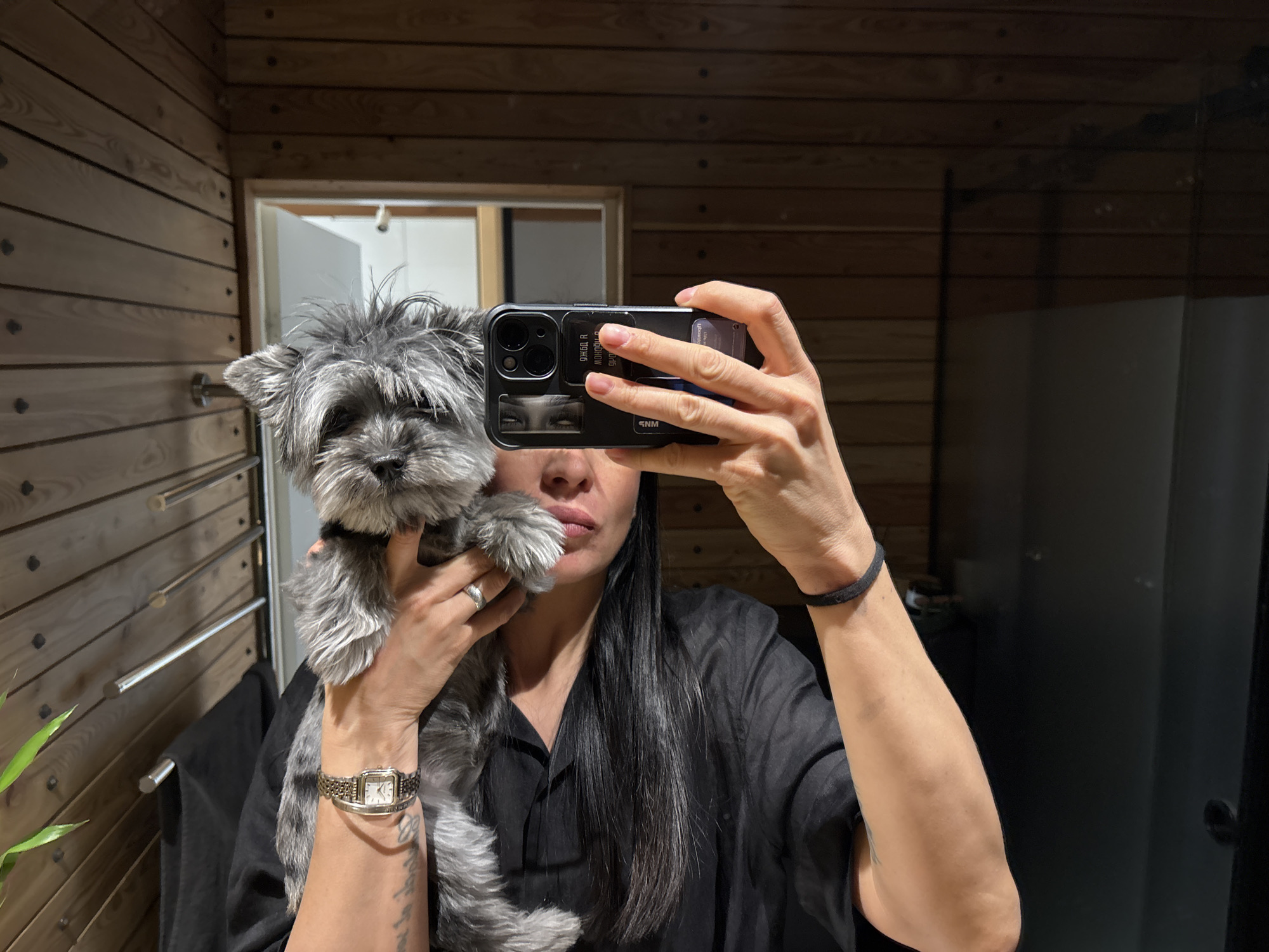 A person is seen in a mirror selfie, holding a small, scruffy-haired grey dog indoors, possibly in Chaikovsky, Russia. The person's face is largely obscured by the smartphone, which they are using to take the picture. They have long, dark hair and are wearing a black shirt. On their left wrist, a square-faced watch with a metal band and a simple bracelet are visible, along with a silver-toned ring on their fourth finger. A black elastic hair tie is on their right wrist. The smartphone appears to be an iPhone in a black case, featuring decorative stickers on the back, including a pair of eyes and reversed text that reads "Я ОДНА В МОНОПОЛИИ" (I AM ALONE IN MONOPOLY) and "SNM".
The setting is an indoor space characterized by prominent horizontal wooden paneling on the walls, suggesting a rustic bathroom, changing room, or sauna area. Reflected in the mirror behind the person is a partial view of a white door and frame. To the left, a structure with several horizontal metal rods, possibly a towel rack or warmer, is visible, along with a small green plant. A dark, possibly black, piece of furniture or wall is reflected on the right side of the image. The lighting is artificial, creating a warm, enclosed atmosphere. The person appears to be casually posing with their pet.