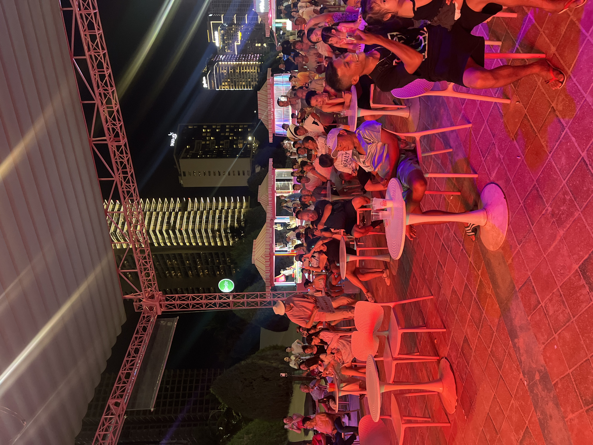 This image depicts an outdoor seating area illuminated by red and pink lights, likely an event space or restaurant in Đà Nẵng, Vietnam. In the background, tall, modern buildings with illuminated windows and a bright green circular sign are visible against a dark night sky, suggesting an urban setting. The foreground and middle ground are occupied by an audience seated at tables and chairs, facing towards an unseen stage or focal point.
The people in the image are diverse in age and gender, dressed in casual attire. Many are looking in the same direction, indicating they are watching a performance or presentation. Some individuals are holding phones, possibly recording or taking pictures. There are several white tables and chairs scattered throughout the scene. A pink truss structure is overhead, supporting a white canopy that provides shade.
In the middle ground, there are what appear to be small, open-air stalls or booths with striped awnings, hinting at a market or food vendors. The ground is paved with reddish-brown tiles. The overall atmosphere appears to be lively and social, with a gathering of people enjoying an evening event.