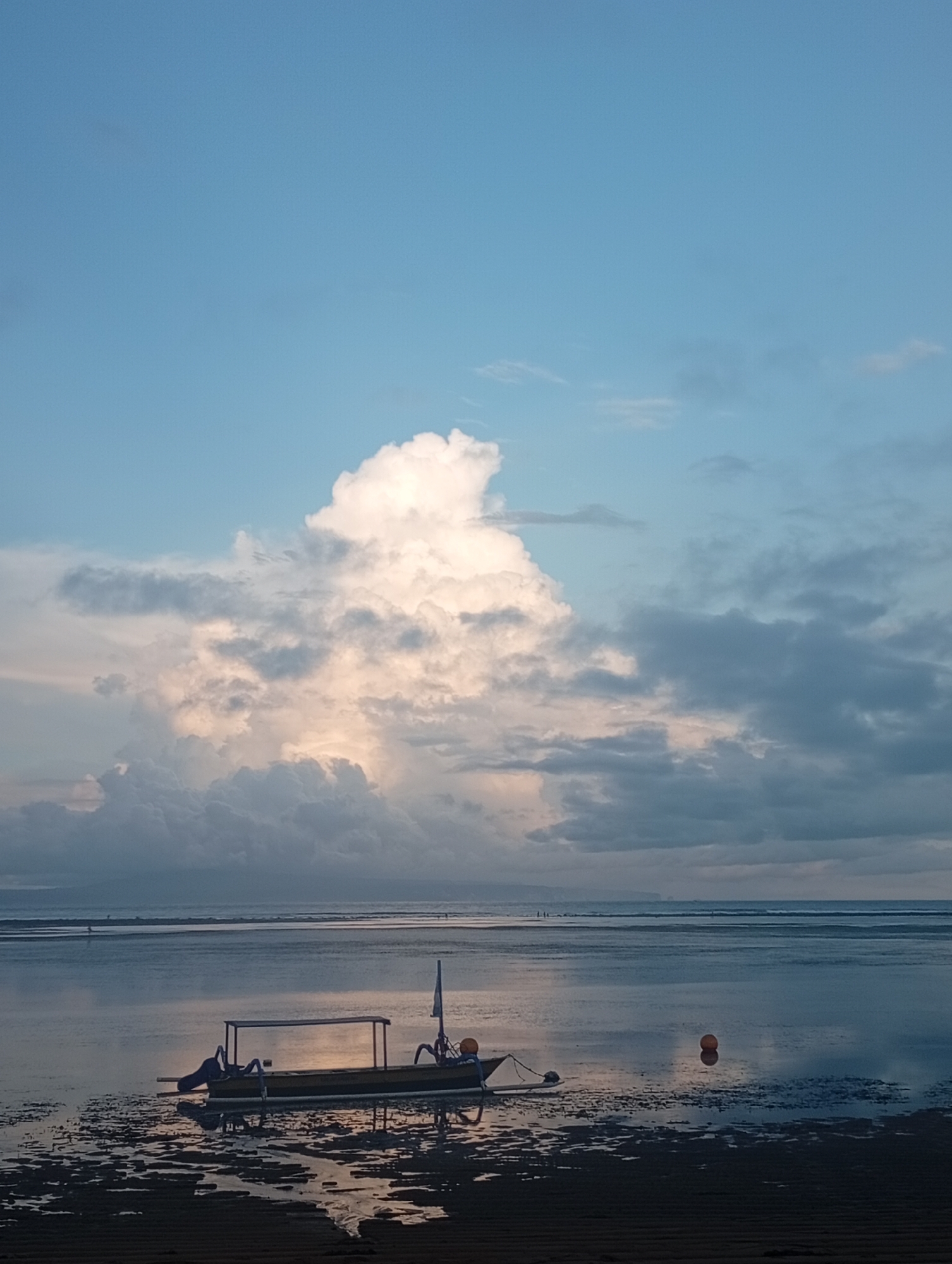The image depicts a coastal scene, likely at low tide, in or near Denpasar, Indonesia, judging by the traditional outrigger boat present. The foreground consists of a dark, wet sandflat with shallow pools reflecting the sky. A traditional wooden boat rests on the exposed seabed, its dark silhouette contrasting with the reflective water. The boat has a simple structure, possibly with a rudimentary shelter. In the middle ground, the calm water stretches out to the horizon, with distant figures appearing as small silhouettes on the waterline. An orange buoy is visible near the boat. The sky is predominantly blue, with a large, towering cumulonimbus cloud formation dominating the center. The clouds range in color from bright white where the sun illuminates them to darker gray shades further out. The overall scene suggests a tranquil, potentially early morning or late afternoon, moment with a blend of natural beauty and elements of local fishing culture.